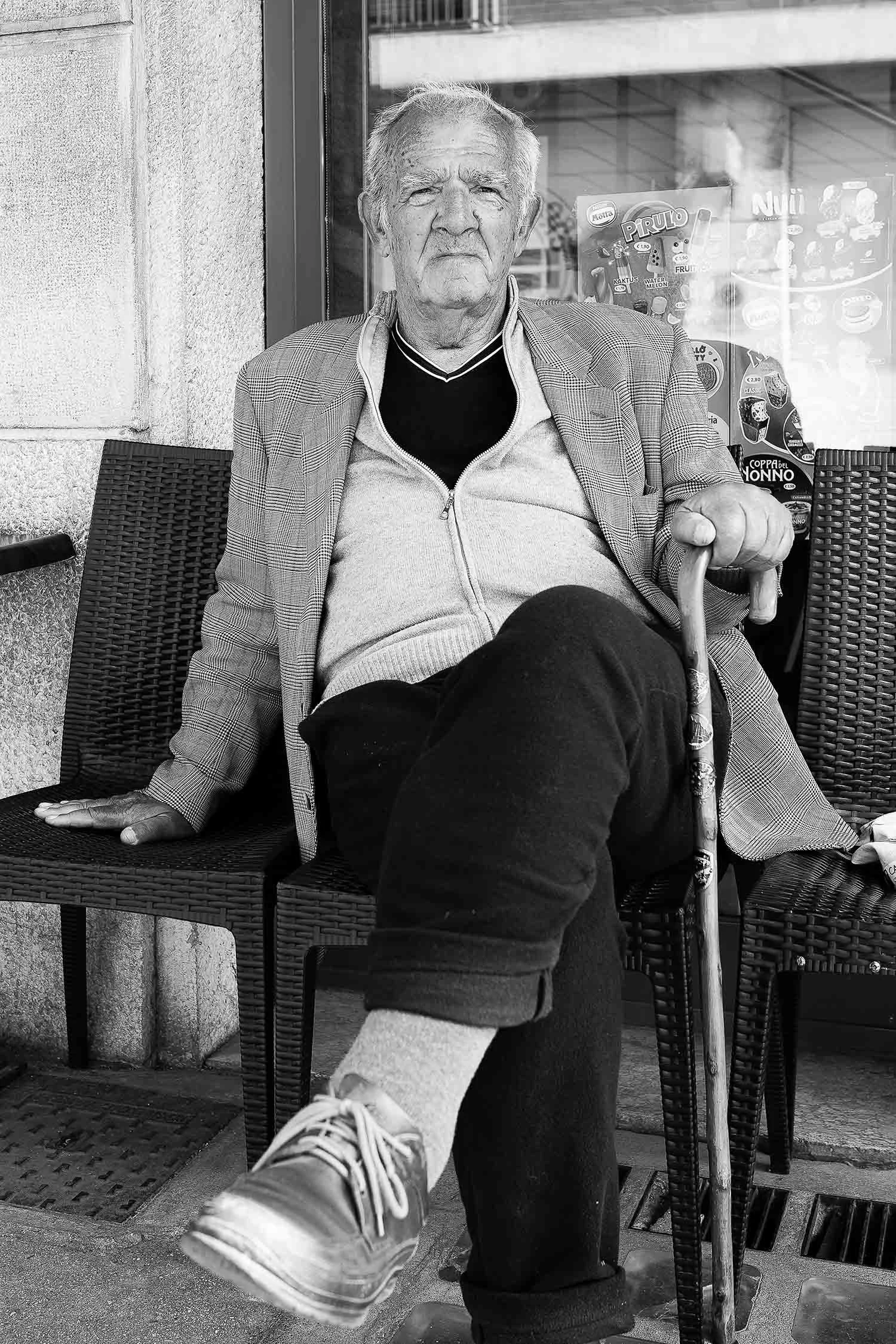 An elderly man sits on a woven chair, dressed in casual layers, holding a cane beside him in a monochrome setting.