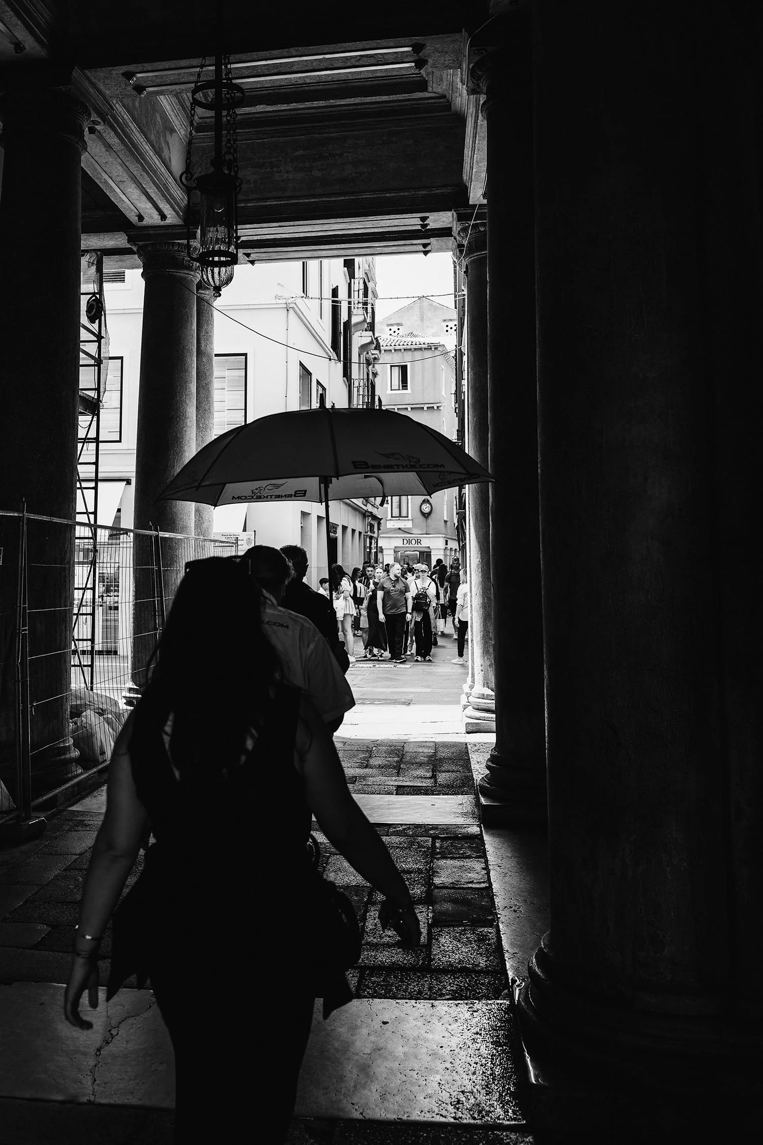 Black and white photo of people walking through a stone-columned archway toward a busy street with umbrellas.