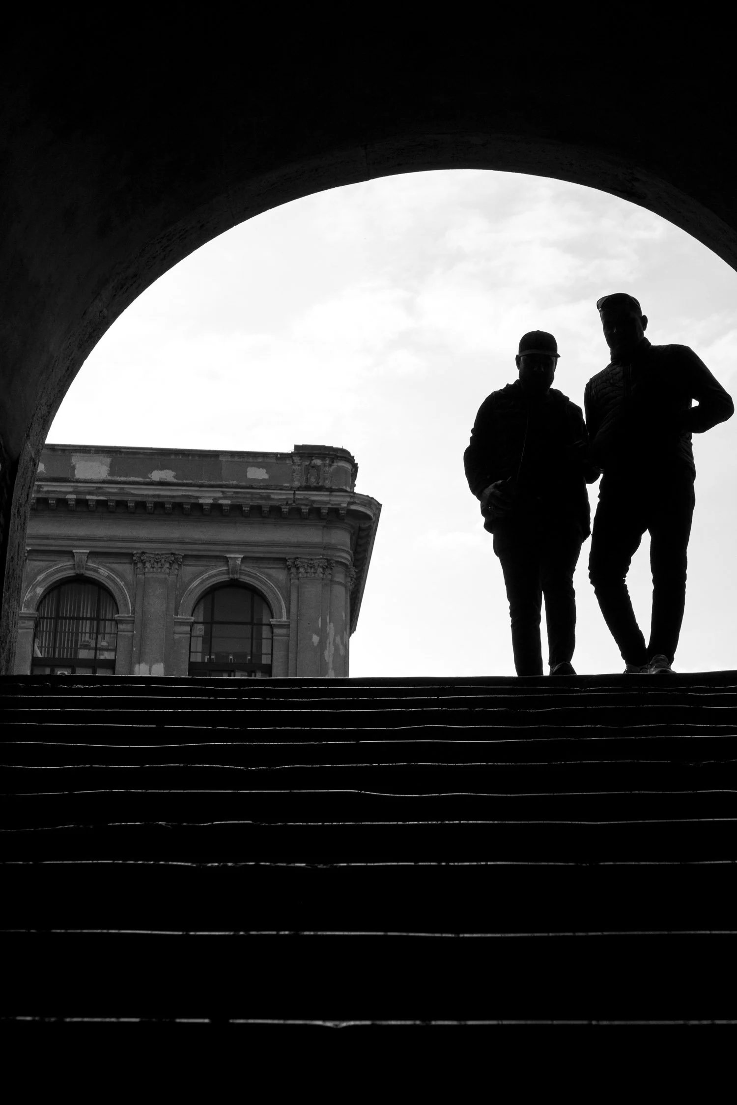 Silhouetted figures stand at the top of a staircase, framed by an arch, with a classic building in the background under a cloudy sky.