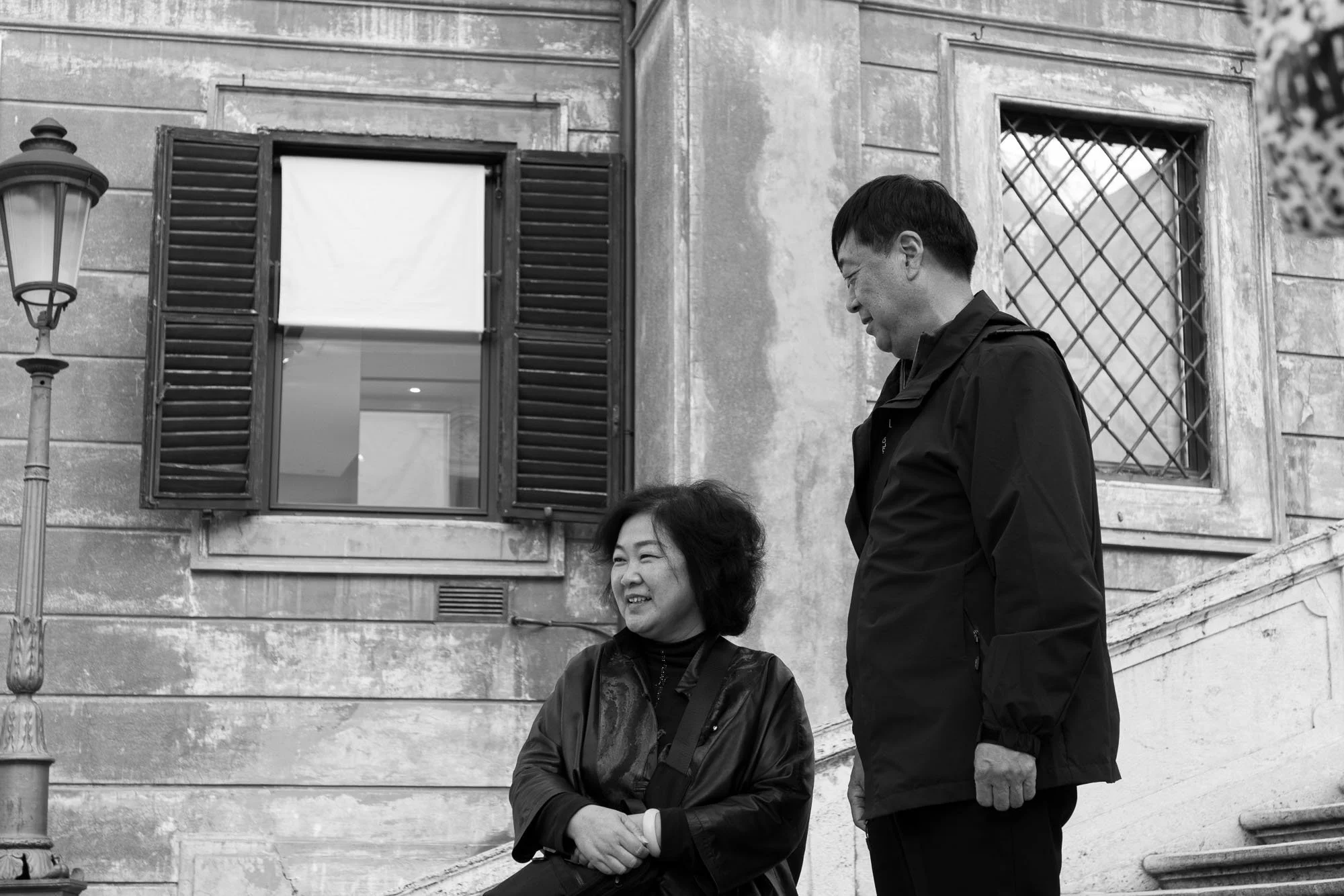 A couple stands on stone steps beside a building with open shutters, deep in conversation, captured in black and white.