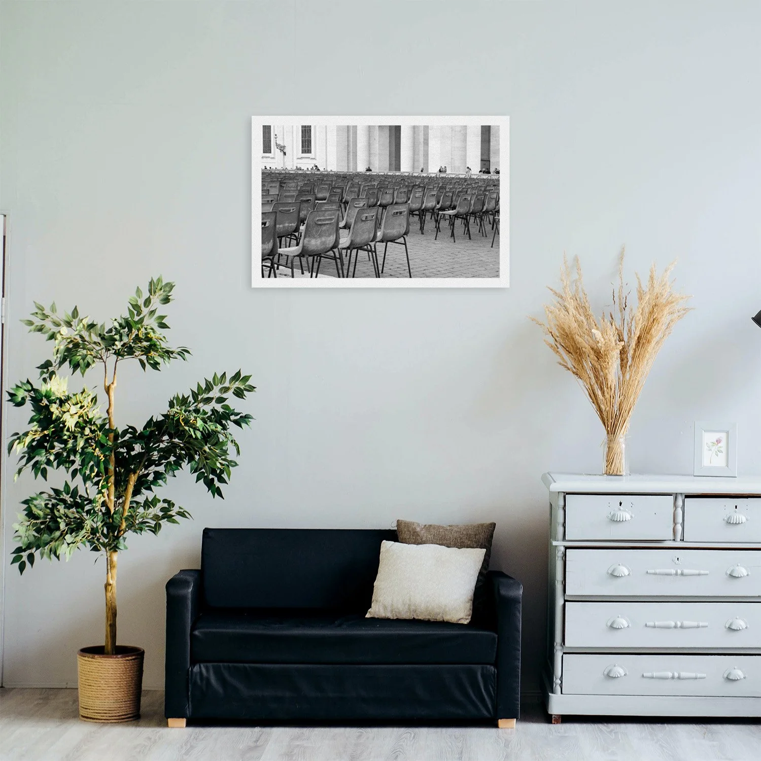 A room interior featuring a black sofa, potted tree, light gray dresser, and framed black and white photo of empty rows of chairs outside a classical building.