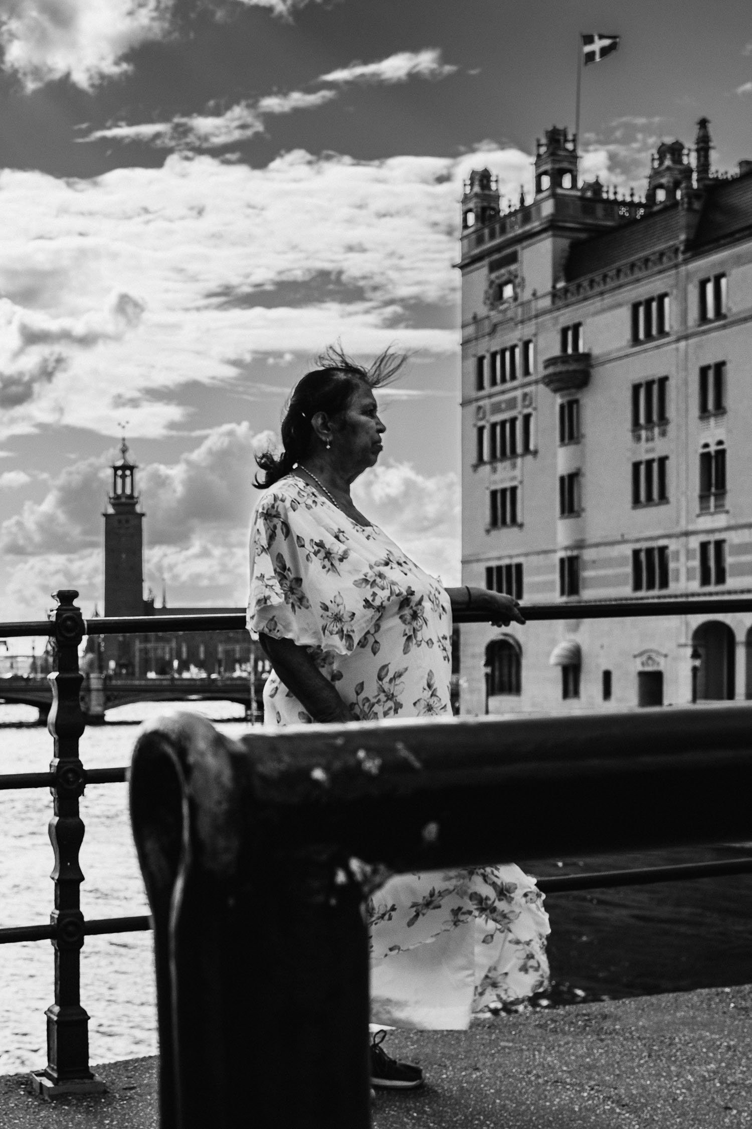 Black and white photo of a woman in a floral dress standing by a waterfront railing with historic buildings and a flag in the background.