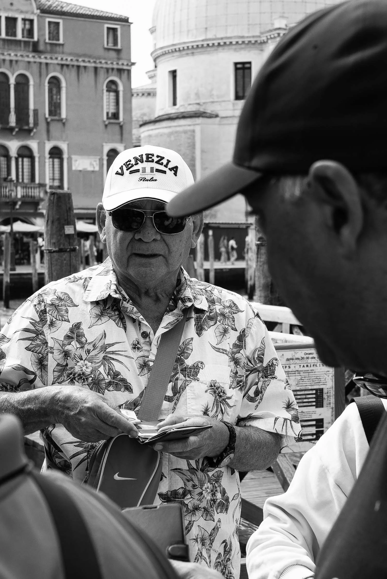 A man wearing a floral shirt and a "VENEZIA" cap exchanges currency at a bustling Venetian waterfront.