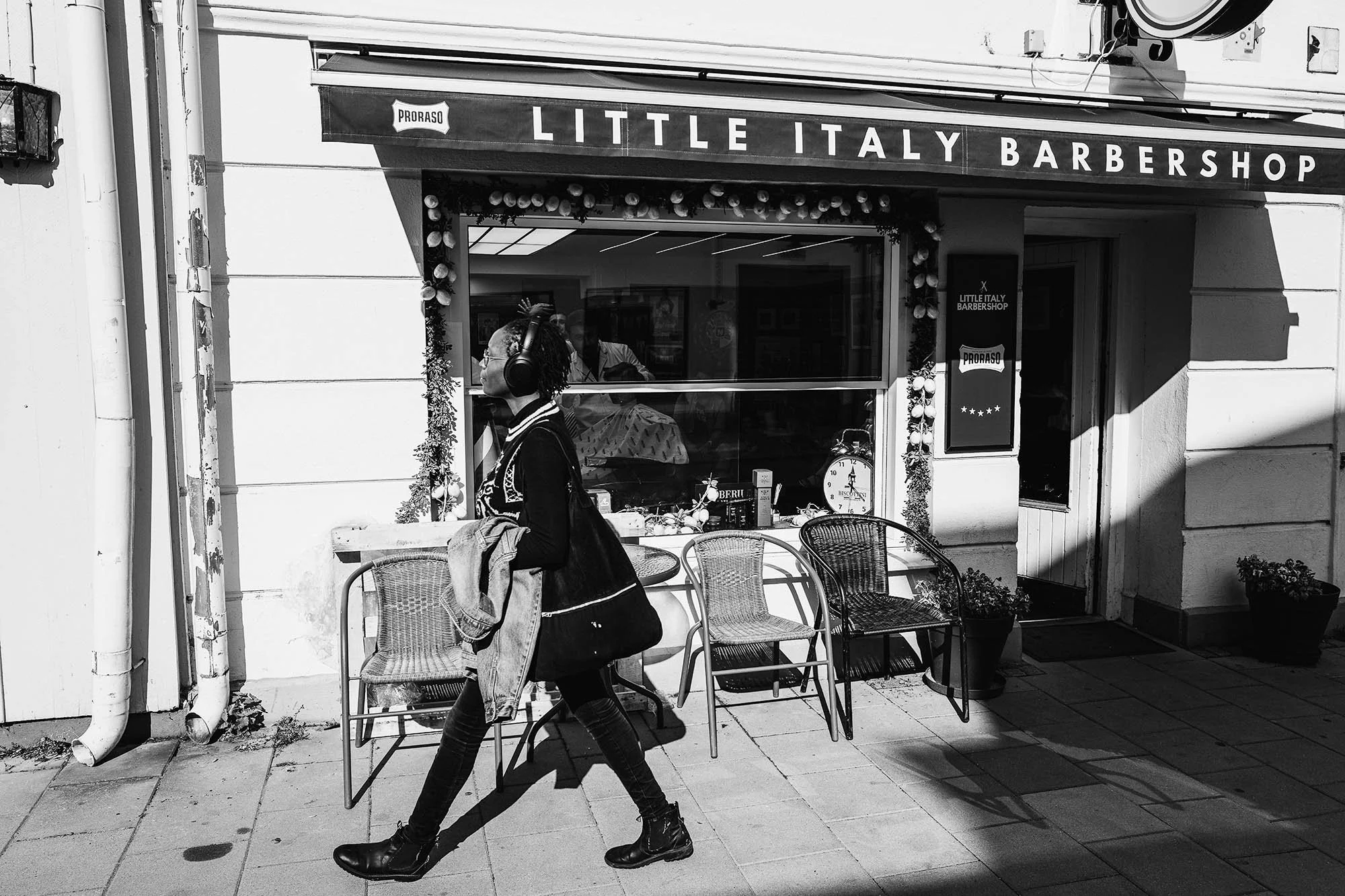 Black-and-white photo of a person walking past Little Italy Barbershop with three empty chairs outside on a sunny day.
