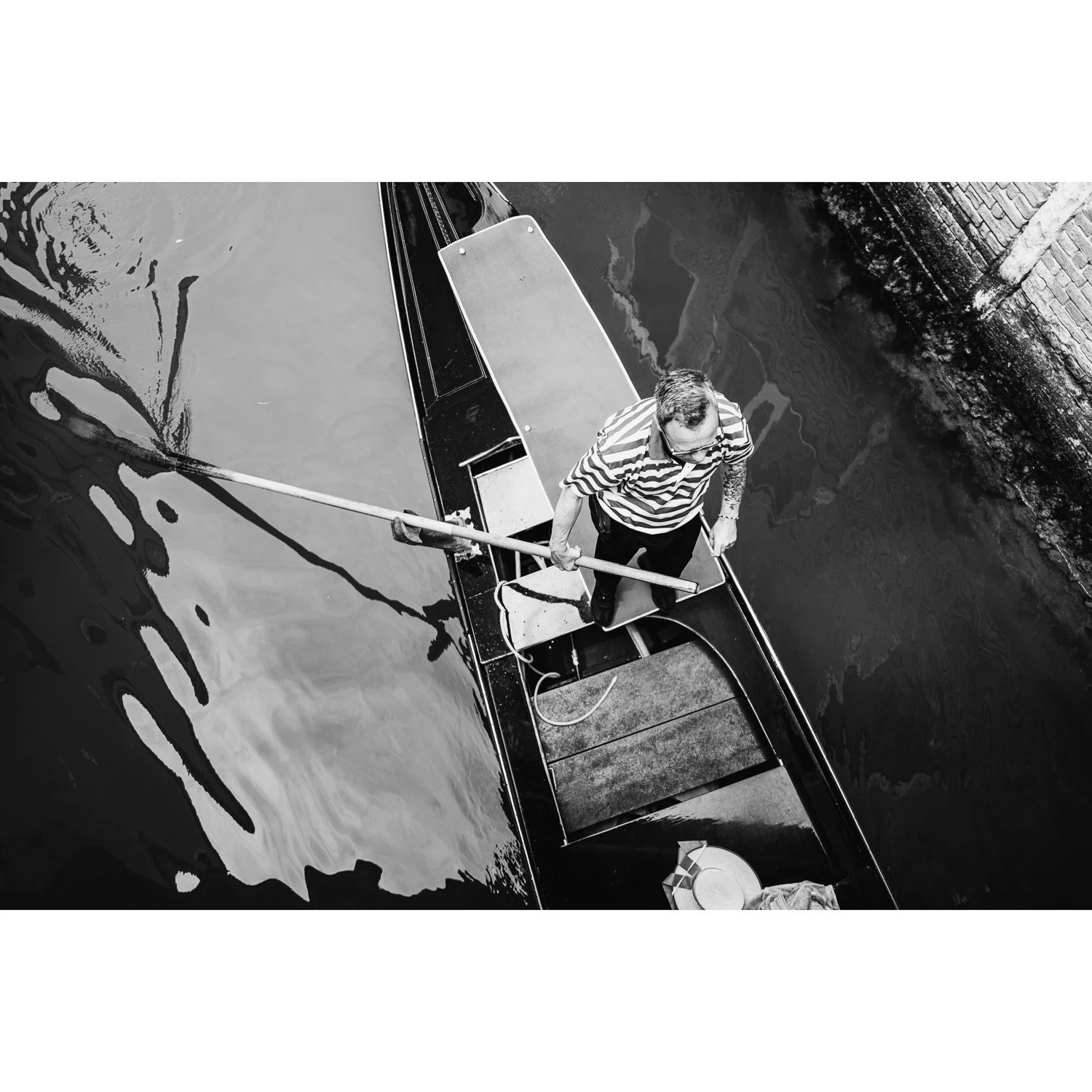 Black-and-white overhead view of a man rowing a narrow boat along a canal.
