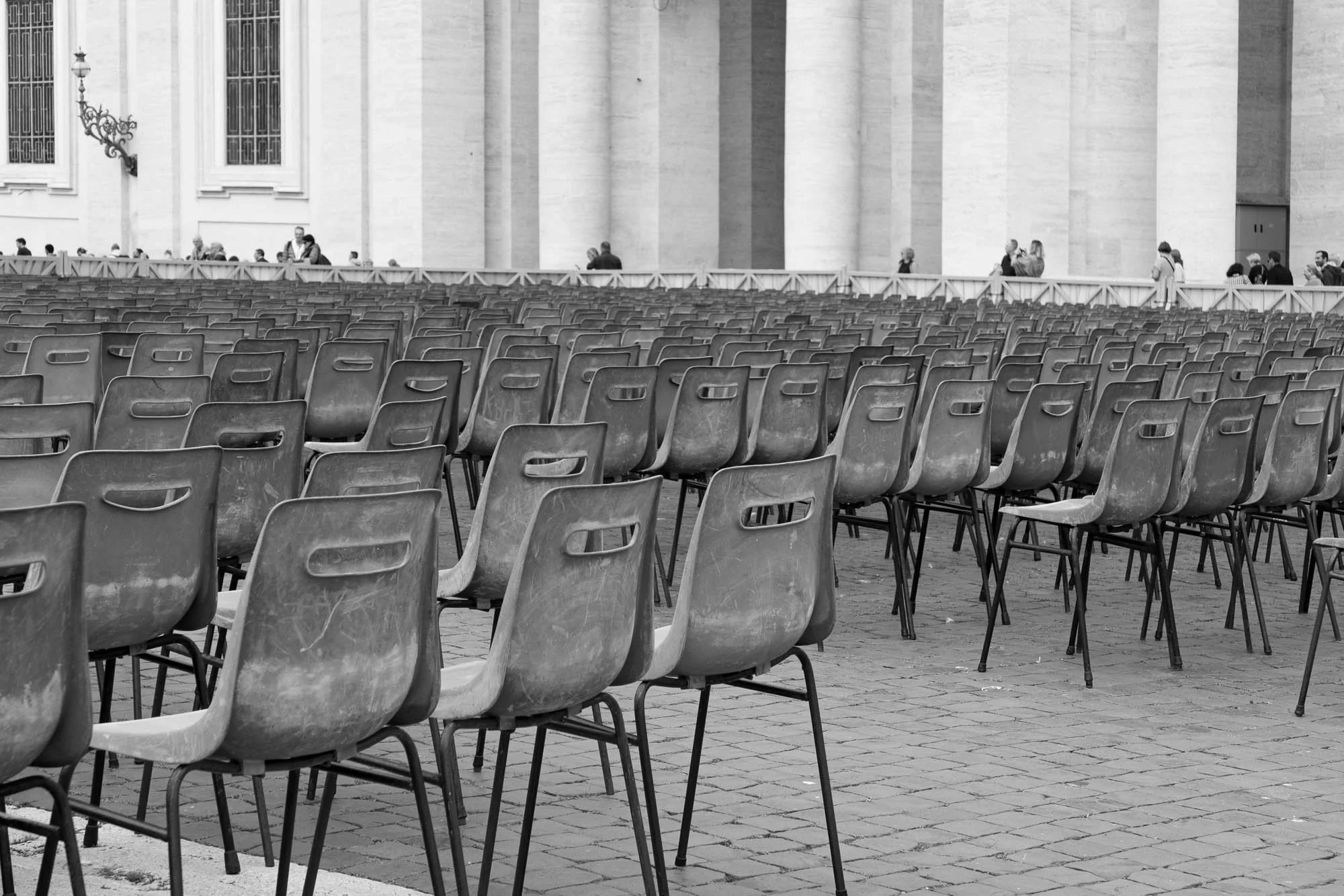 Black and white image depicting empty chairs arranged in front of a building, evoking a sense of solitude and stillness.