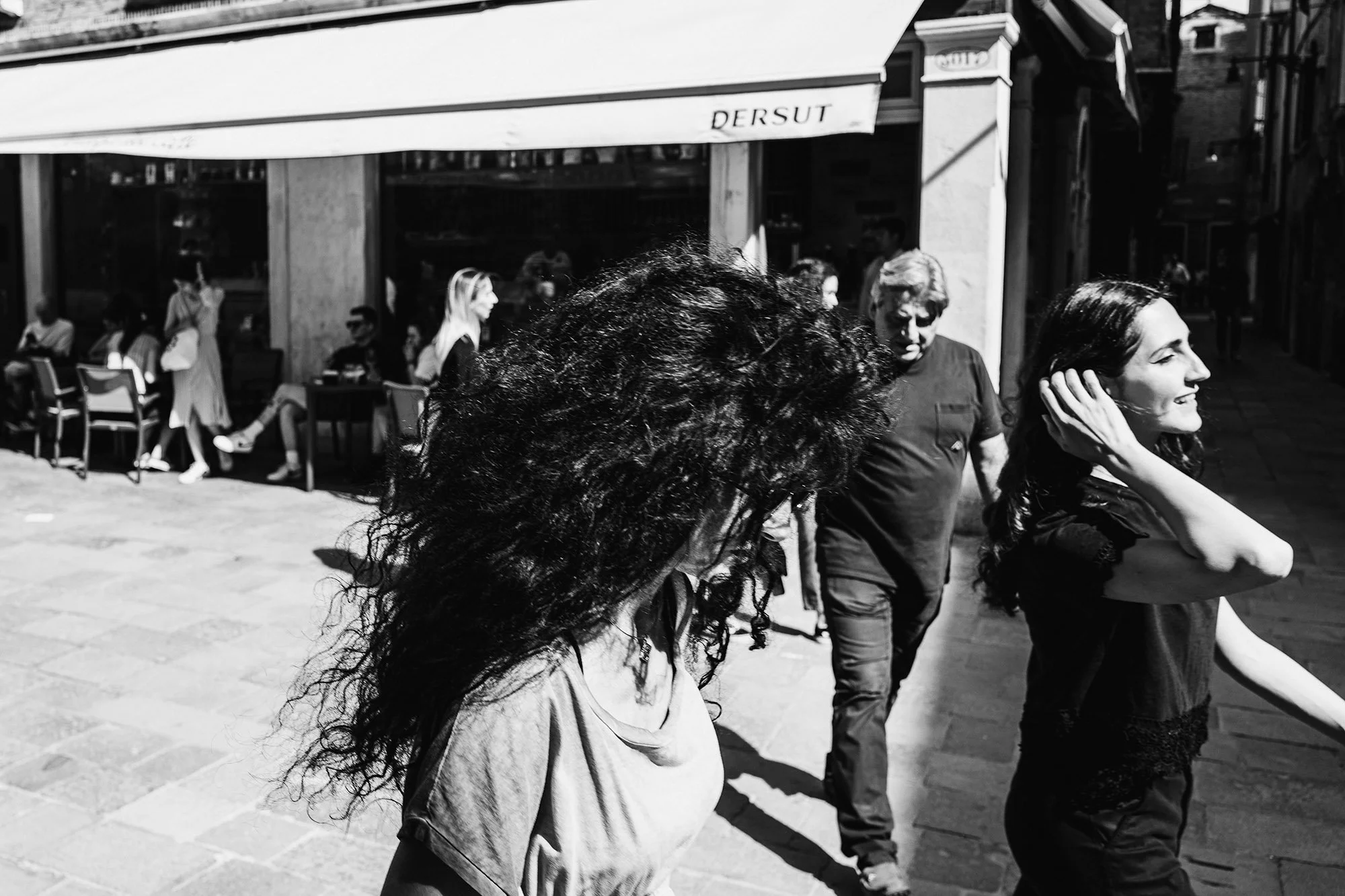 A monochrome street scene featuring two women with long, dark hair walking past a man, surrounded by urban architecture and outdoor seating.
