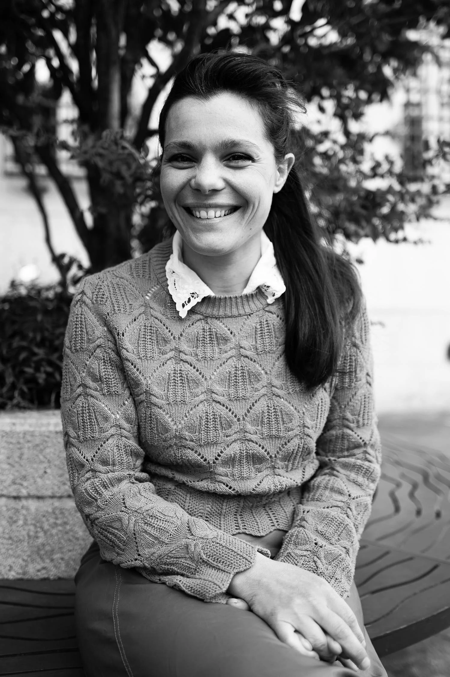 A woman sits outdoors, wearing a knitted sweater and a white collared shirt, with a natural backdrop of greenery and textured pavement.