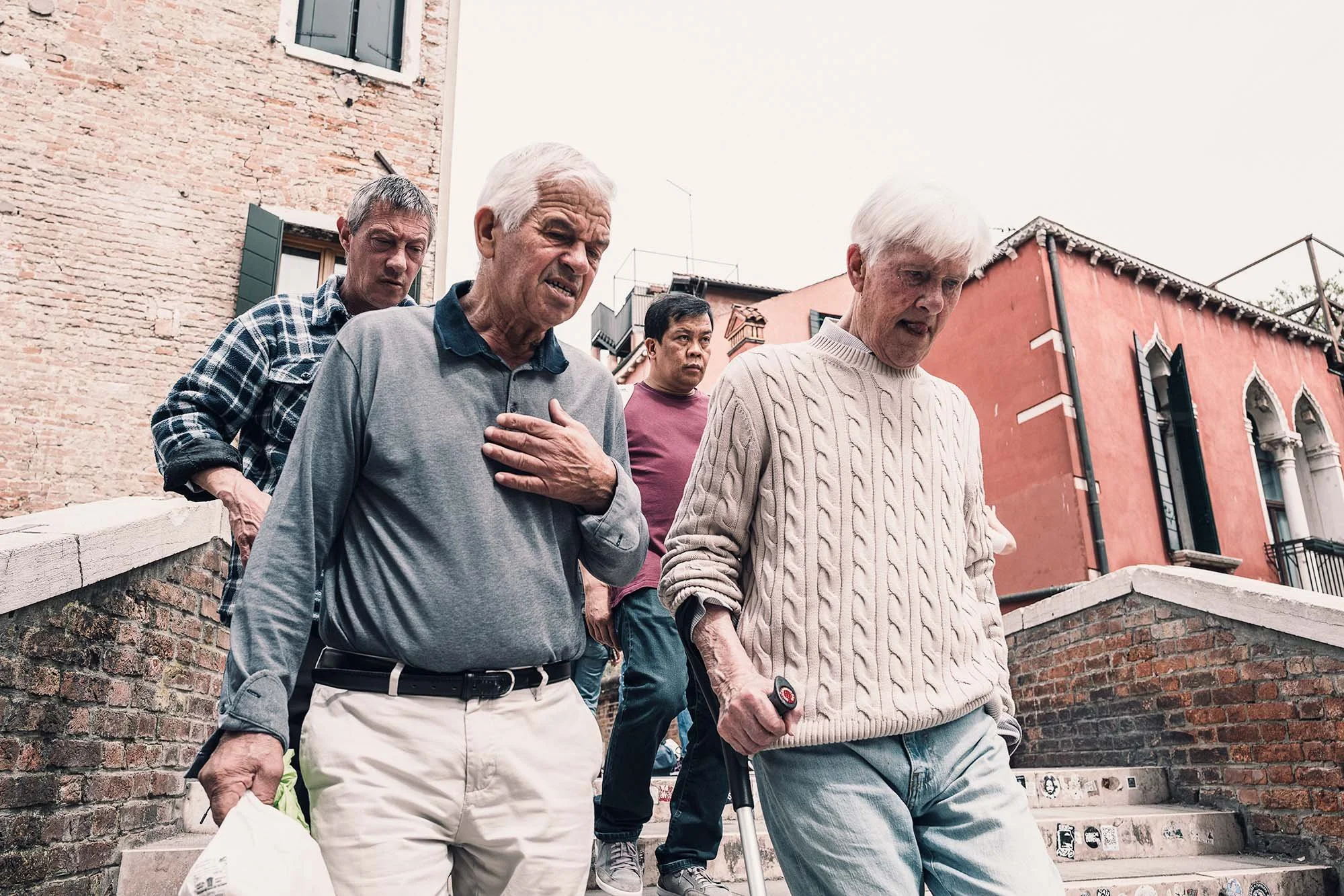 Four elderly men walking down outdoor steps in a historic urban area with brick and plaster buildings.