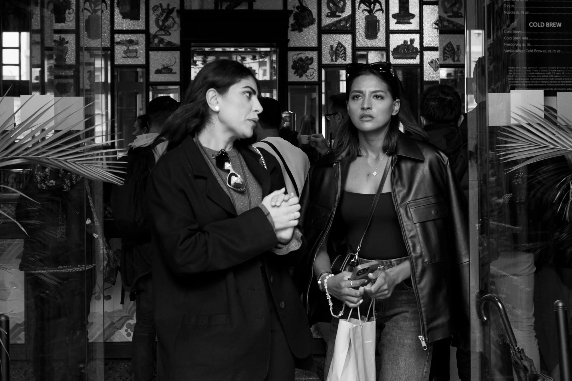 Two women with stylish outfits, standing and talking outside a shop with decorative patterned glass, holding shopping bags.