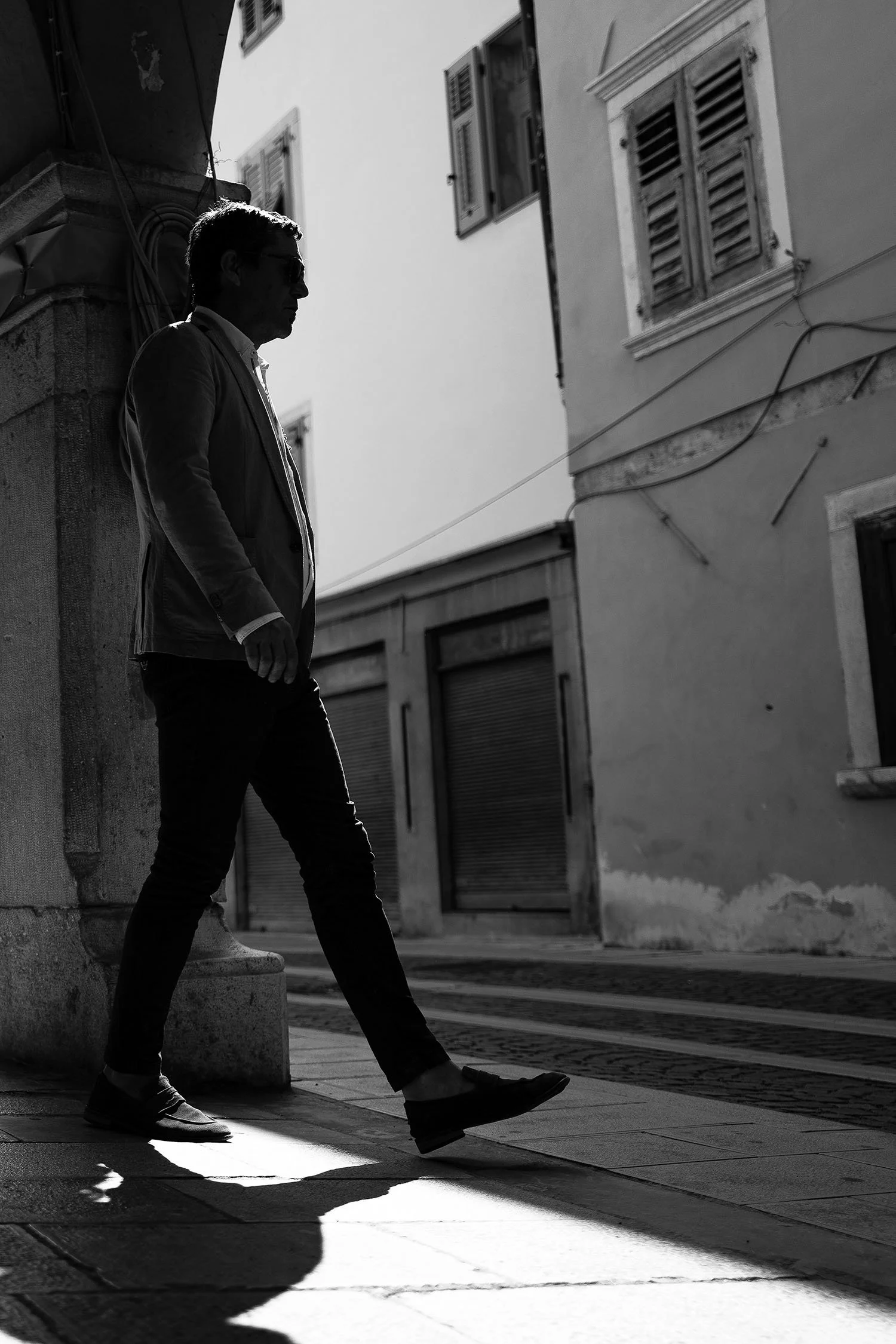 A person in a suit walks down a city street, sunlight casting shadows on the pavement.