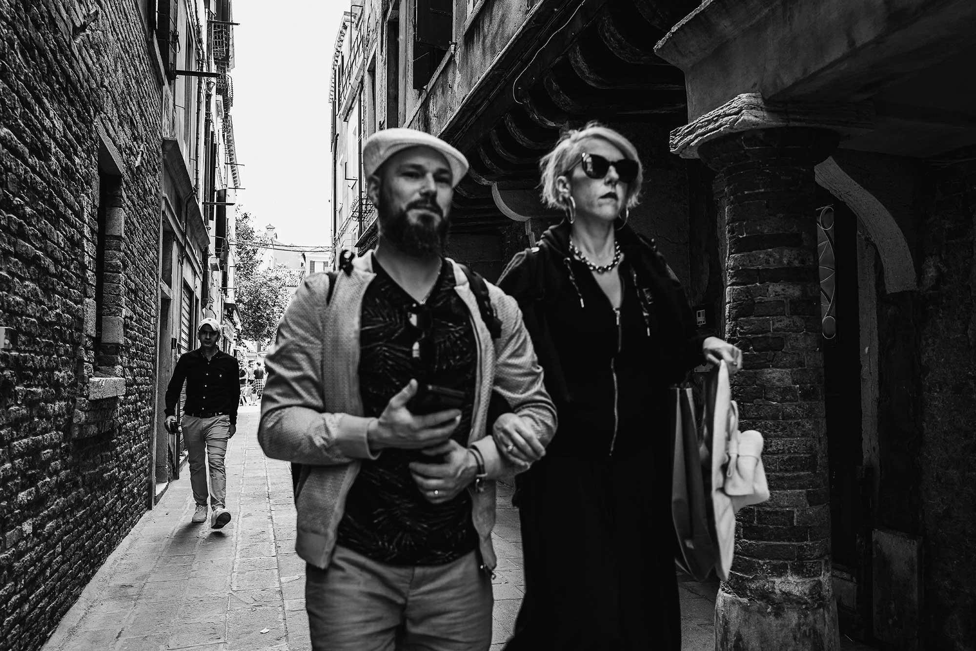 Black and white photo of two people walking arm in arm down a narrow city street with a third person behind them.