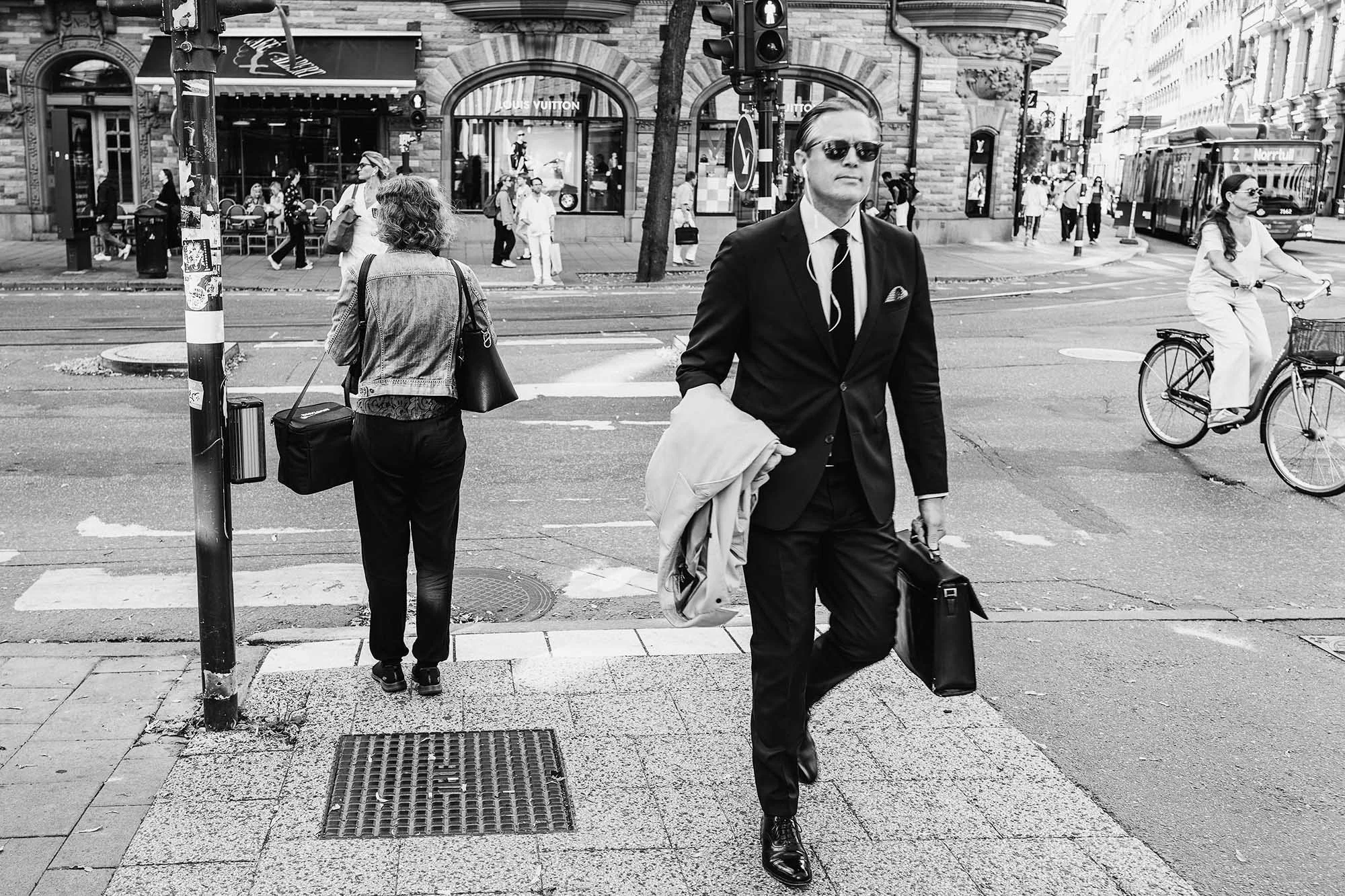 Black and white street scene with a man in a suit holding a briefcase, a woman waiting at a crosswalk, and a cyclist passing by.