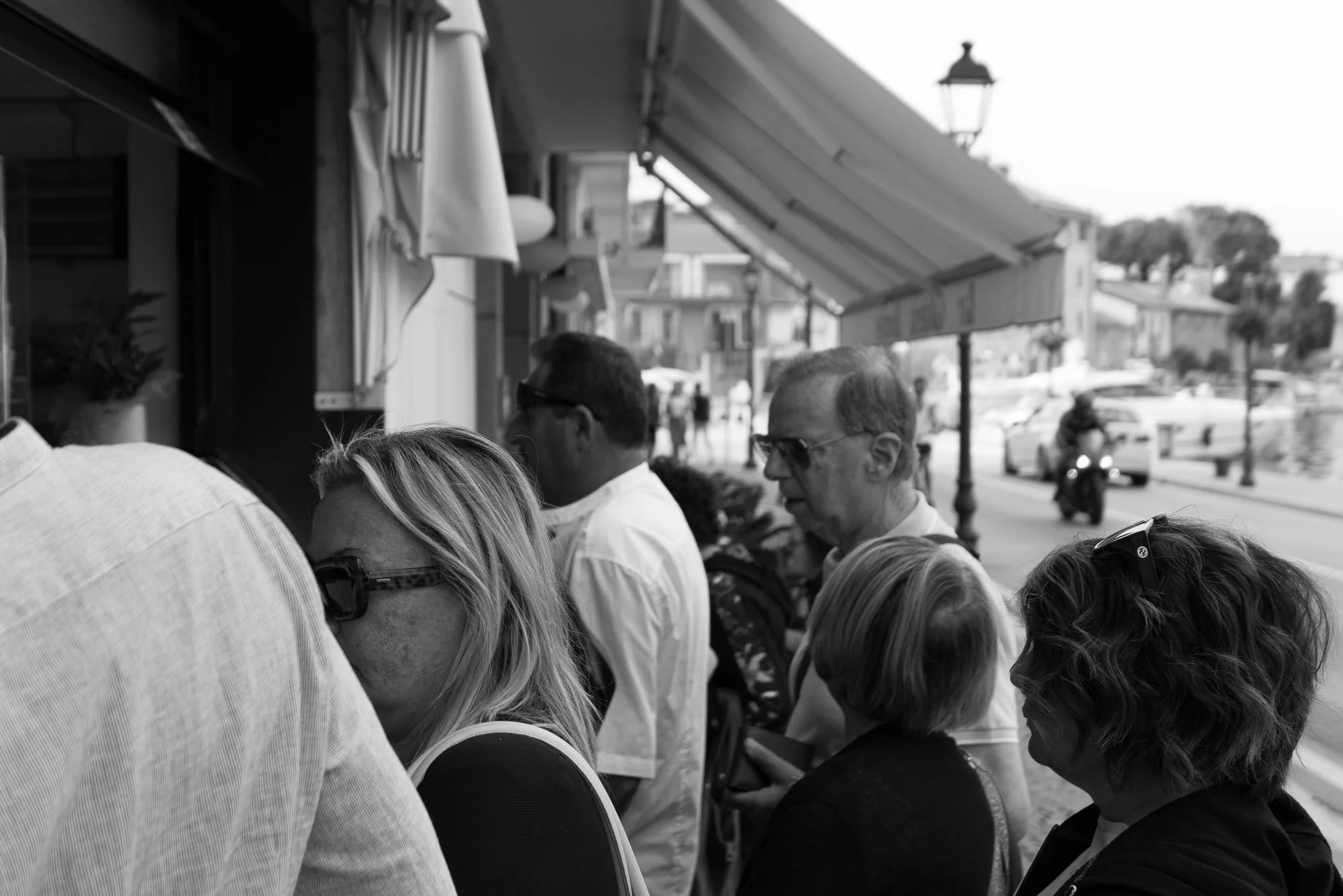 A black and white image of a crowded street, with people waiting in line outside a shop under an awning.