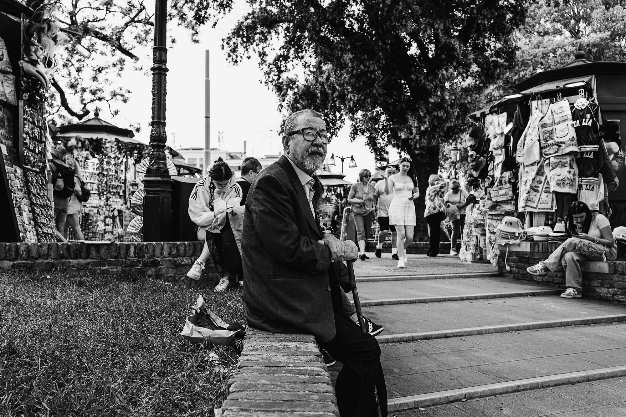 A man sits pensively on a stone bench, surrounded by bustling tourists and colorful stalls in a lively outdoor market.
