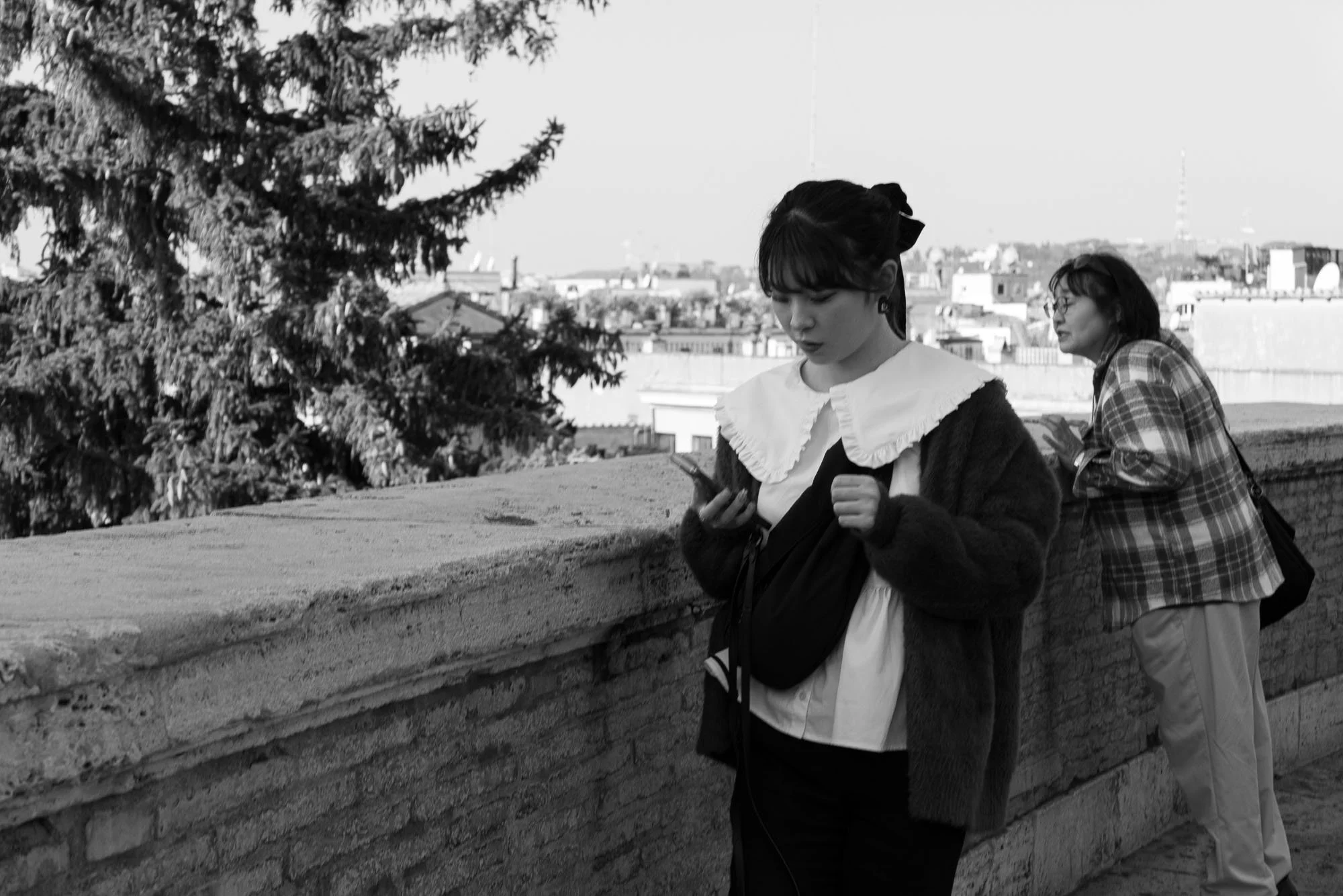 Two women, one looking at a phone, the other observing, stand beside a stone wall with a city view in the background.
