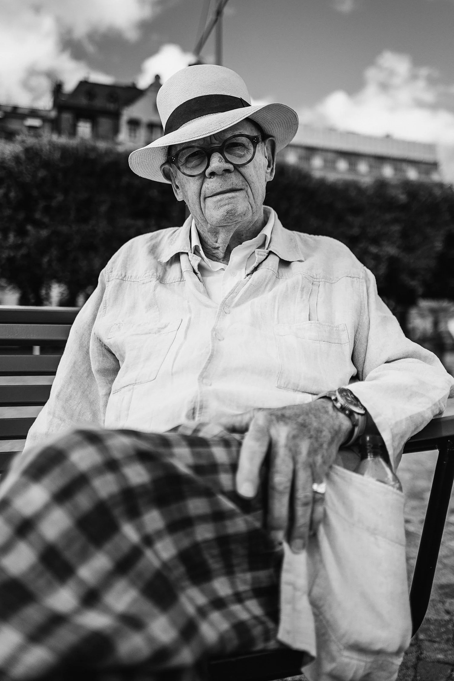 Black and white portrait of an elderly man in a hat and glasses sitting on a bench.