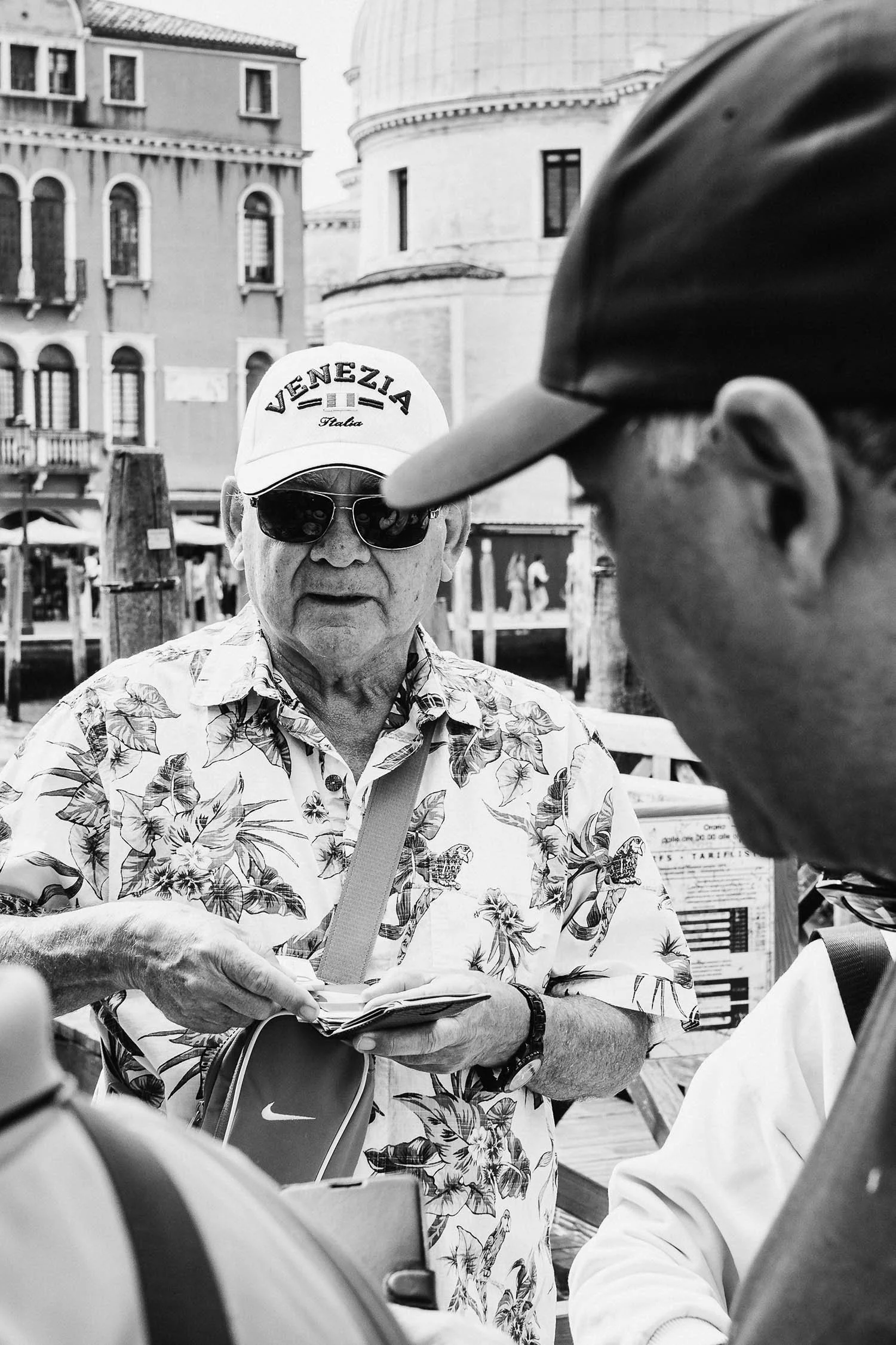 A man wearing a floral shirt and a "VENEZIA" cap exchanges currency at a bustling Venetian waterfront.
