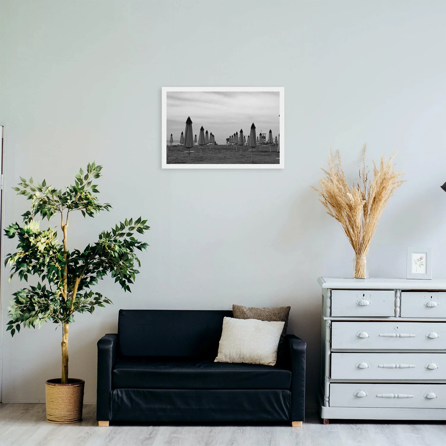 A black and white framed photograph of numerous closed beach umbrellas on a cloudy shore hangs above a black sofa flanked by a potted plant and a light gray dresser with dried grass in a vase.