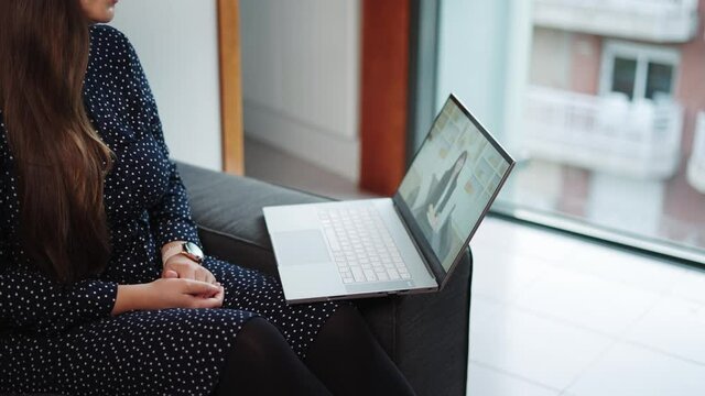 A woman sitting on a gray sofa with a black and white polka dot dress, working on an open laptop near a large window with a city view