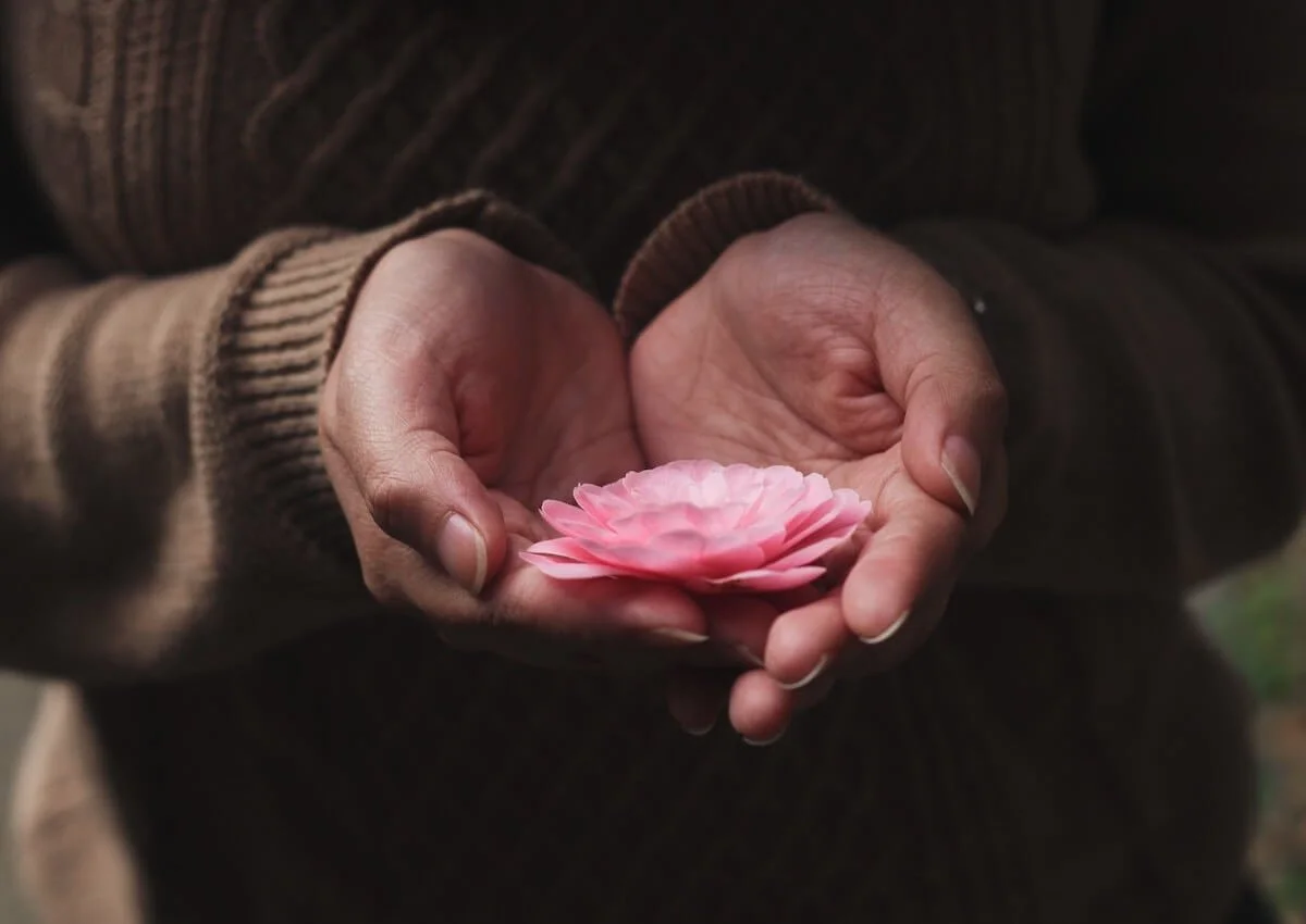 A child holding an adult's finger outdoors near a body of water, with trees in the background, during a cloudy day.