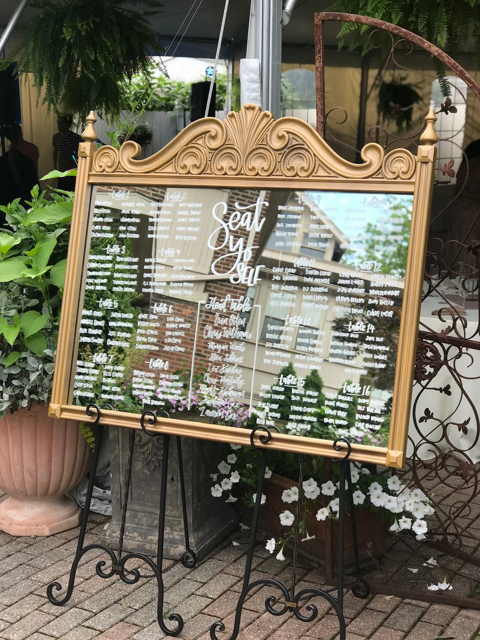 Hand lettered wedding seating chart on a wide, gold, decorative mirror on easels at the entrance of a wedding reception.