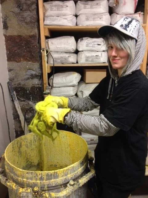 Katie stands in a room holding sculptures above lime green liquid as part of the bronze casting process in school