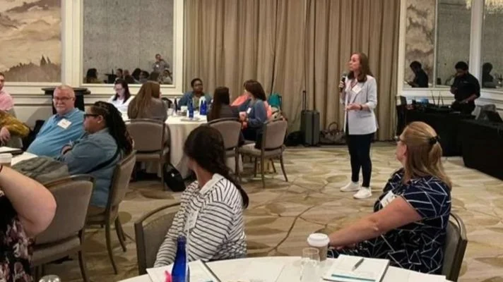A woman is speaking at a conference in a large, decorated room with round tables and seated attendees, some with coffee cups and notebooks, while others listen or look at her.