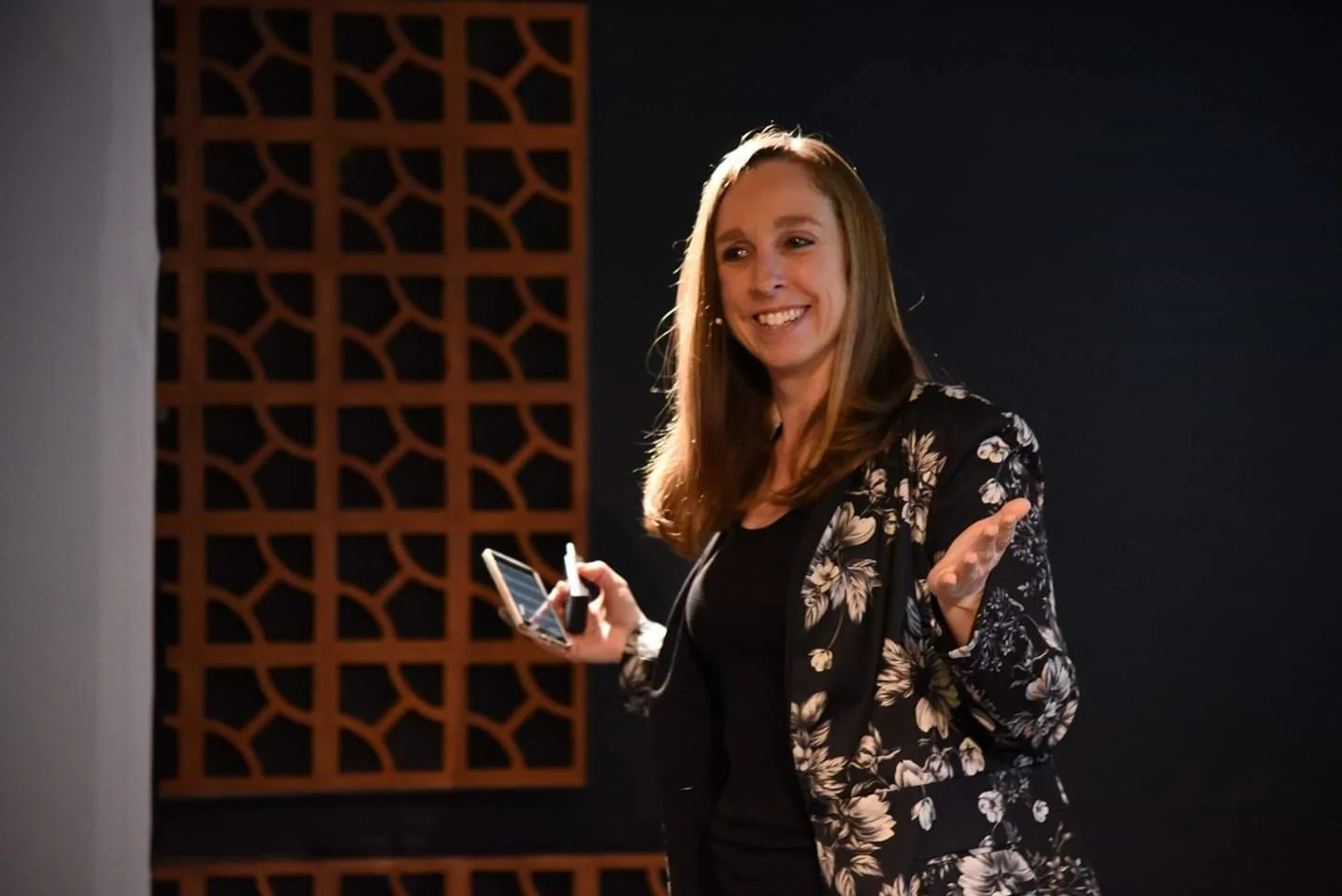 A woman with long brown hair smiling and gesturing with her right hand while holding a smartphone and a marker in her left hand, standing in front of a dark background with a decorative wooden panel.