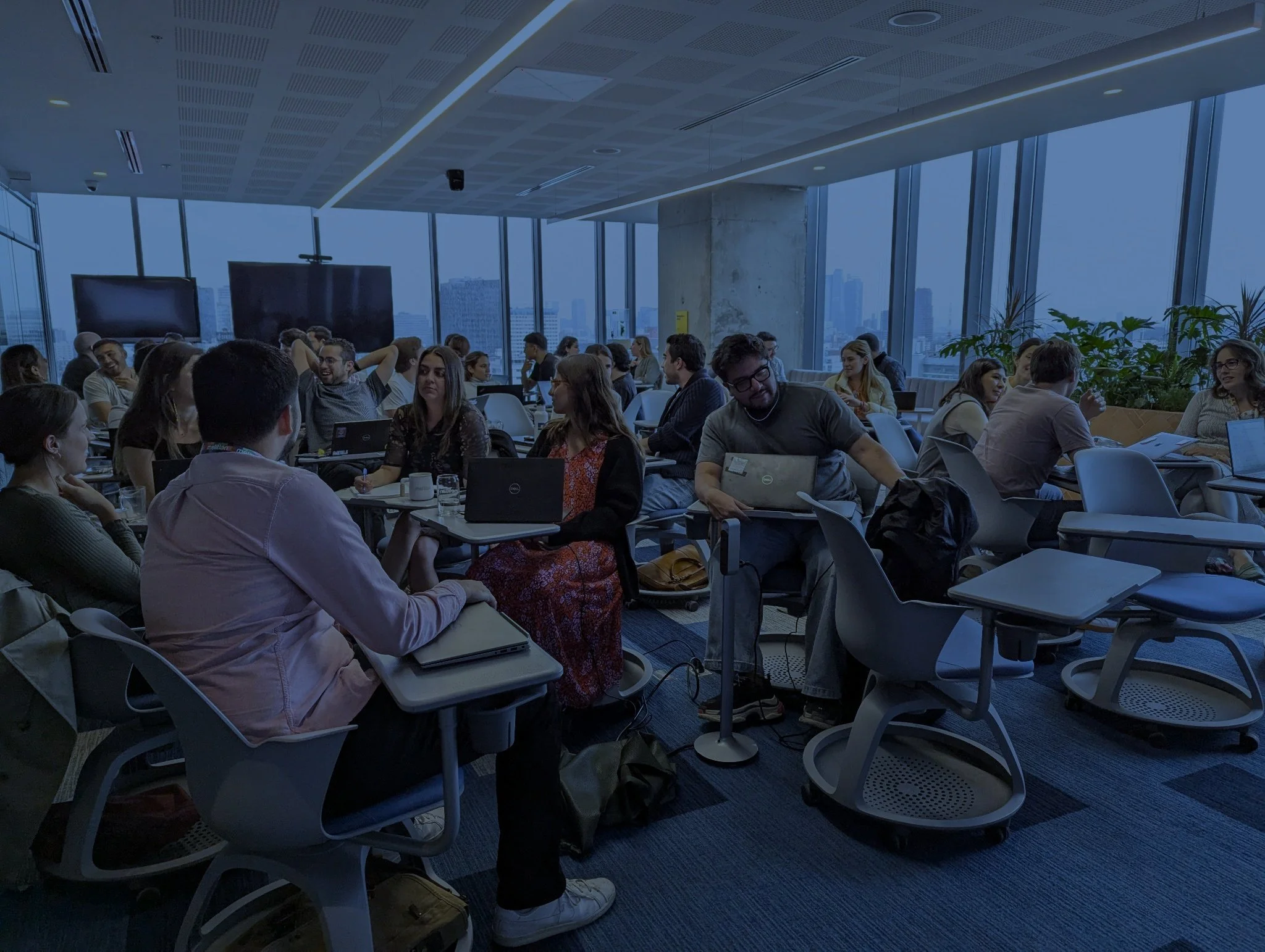 A conference room filled with people of diverse backgrounds sitting at tables with laptops, engaged in discussions and working, with large windows showing a city skyline in the background.