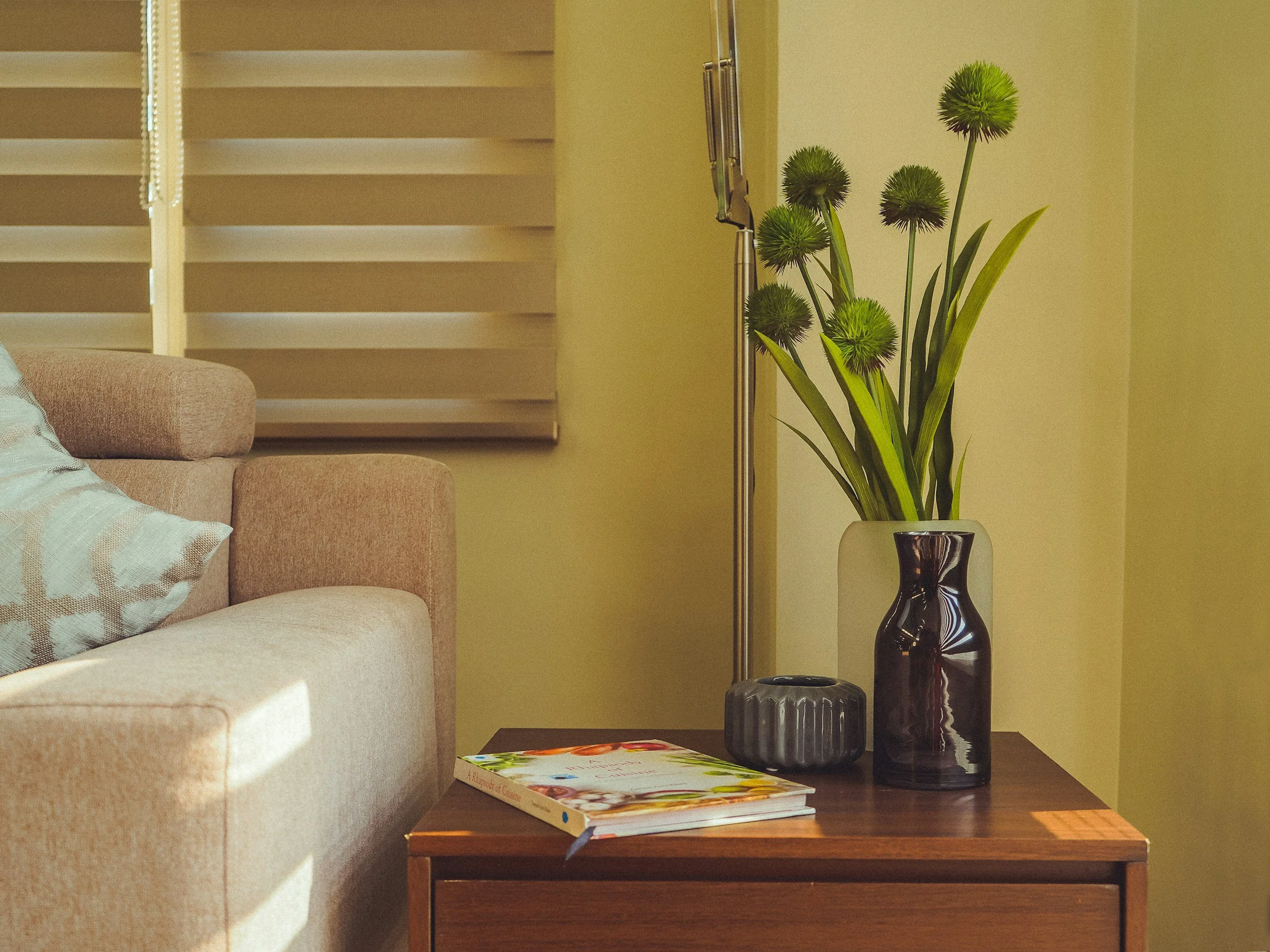 A side table with two dark-colored vases, a stack of colorful books, and a magazine. A beige couch with a patterned pillow is partially visible. In the background, there is a yellow wall, a window with closed blinds, and a white vase with tall green flowers.