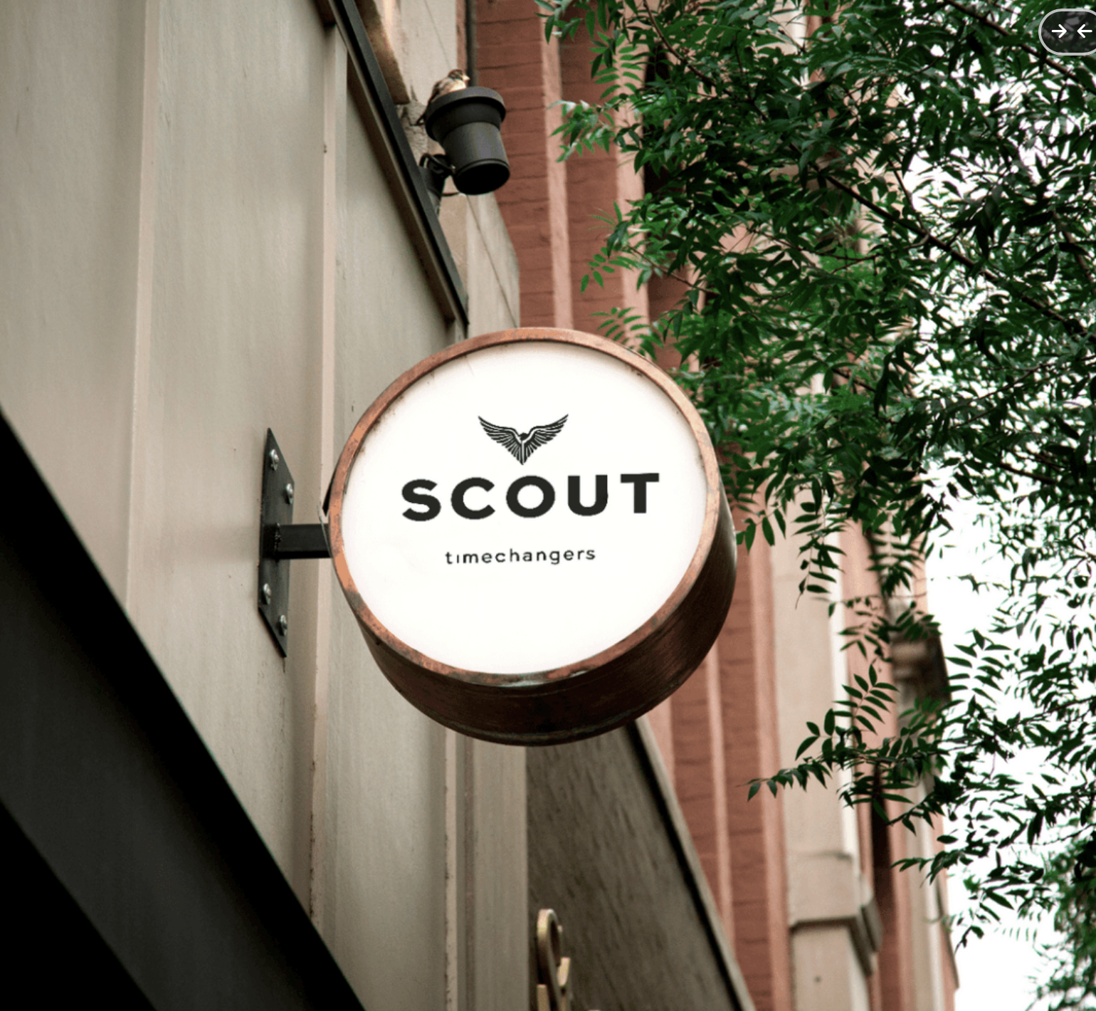 A circular sign for a business called SCOUT, with smaller text 'timechangers', mounted on the side of a building. The sign has a white background, black text, and a winged logo at the top. Trees are visible in the background.