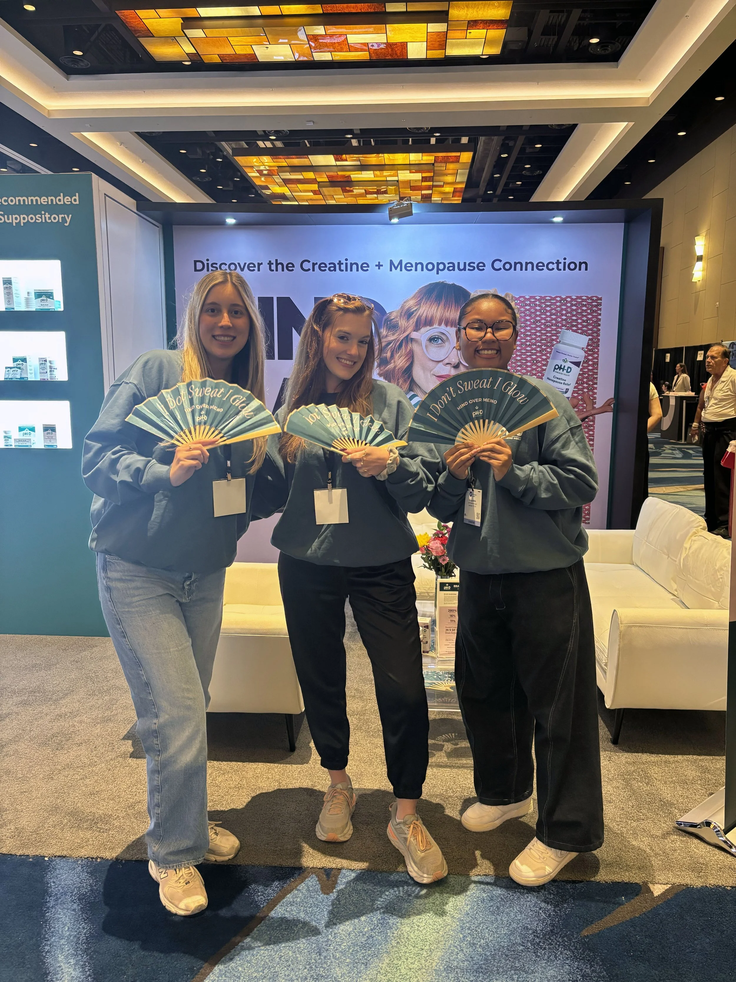 Three women standing together at a conference, holding fans with the text 'I Don't Sweat I Glow'. They are smiling and wearing casual clothing, with a display behind them promoting the connection between creatine and menopause.