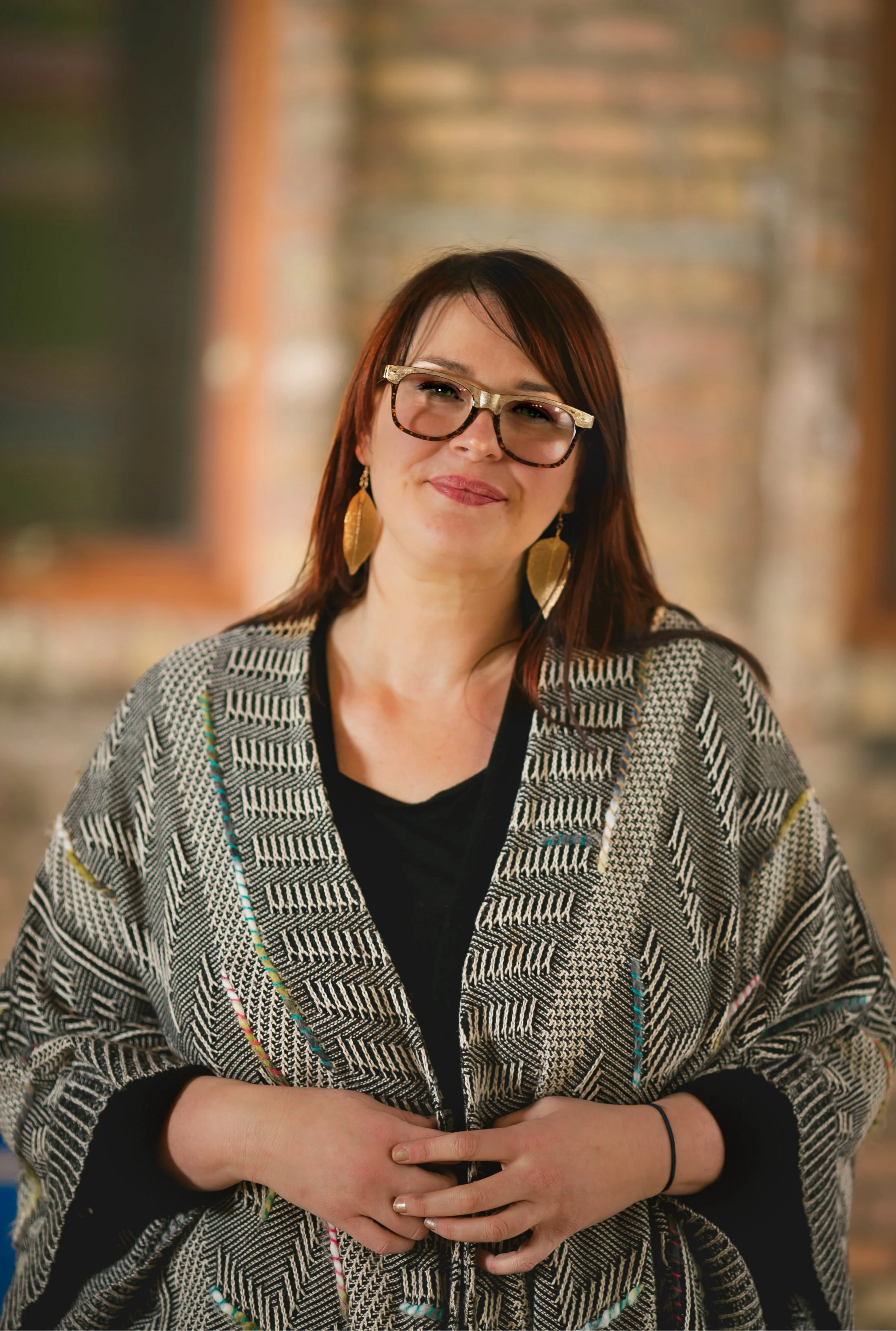 A woman with reddish-brown hair, wearing glasses, gold earrings, and a patterned shawl over a black top, smiling and holding her hands together. Cara Dunning Headshot