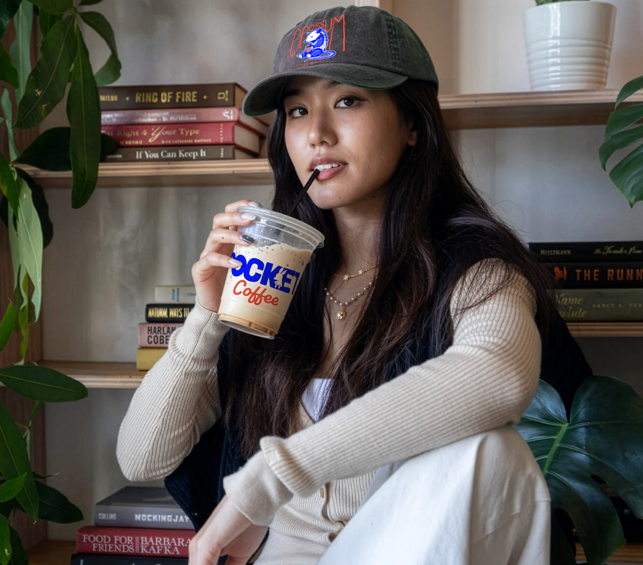 A young woman with long dark hair wearing a baseball cap and beige cardigan sipping iced coffee from a clear plastic cup, sitting in a cozy space with bookshelves and green plants in the background.