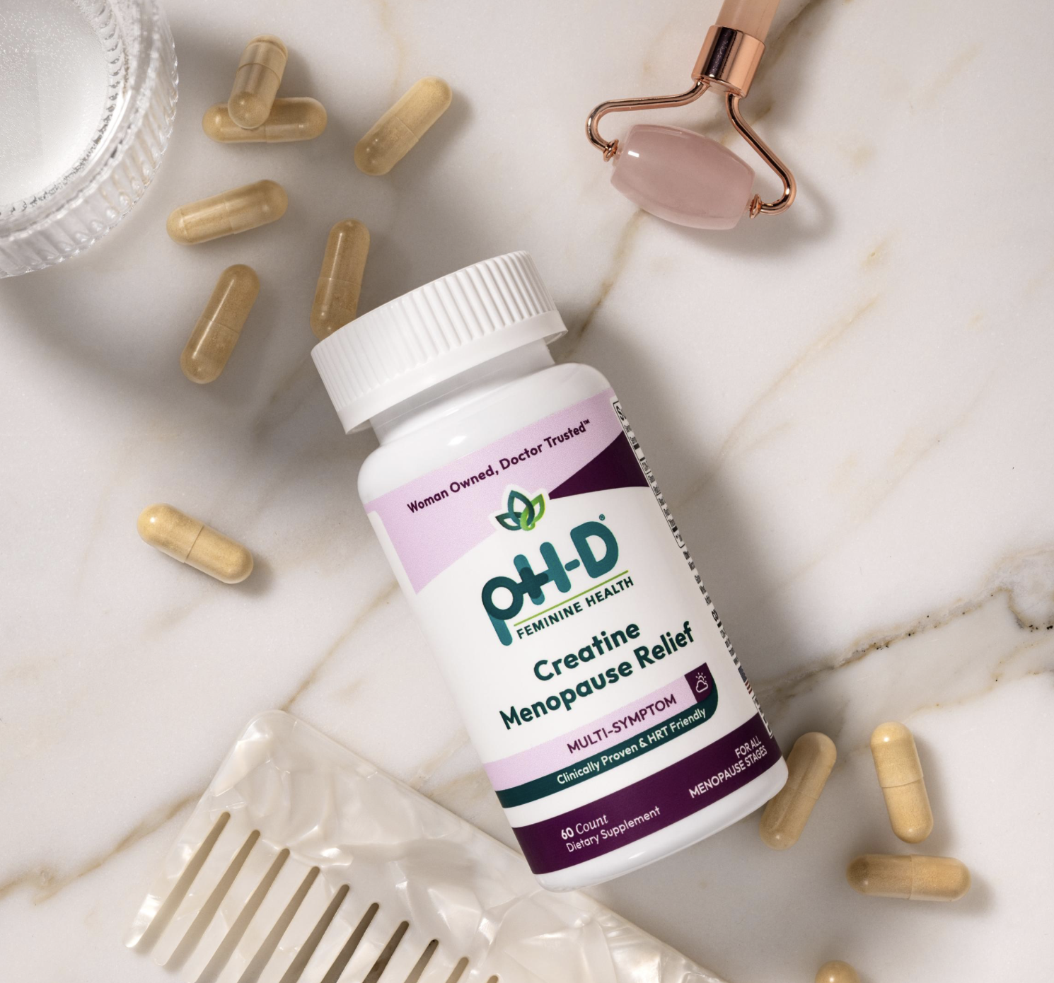 A bottle of pH-D Feminine Health Creatine Menopause Relief supplements surrounded by capsules, a roller massager, and a hair comb on a marble surface. Cara Dunning Art Direction Eric Yu Photographer