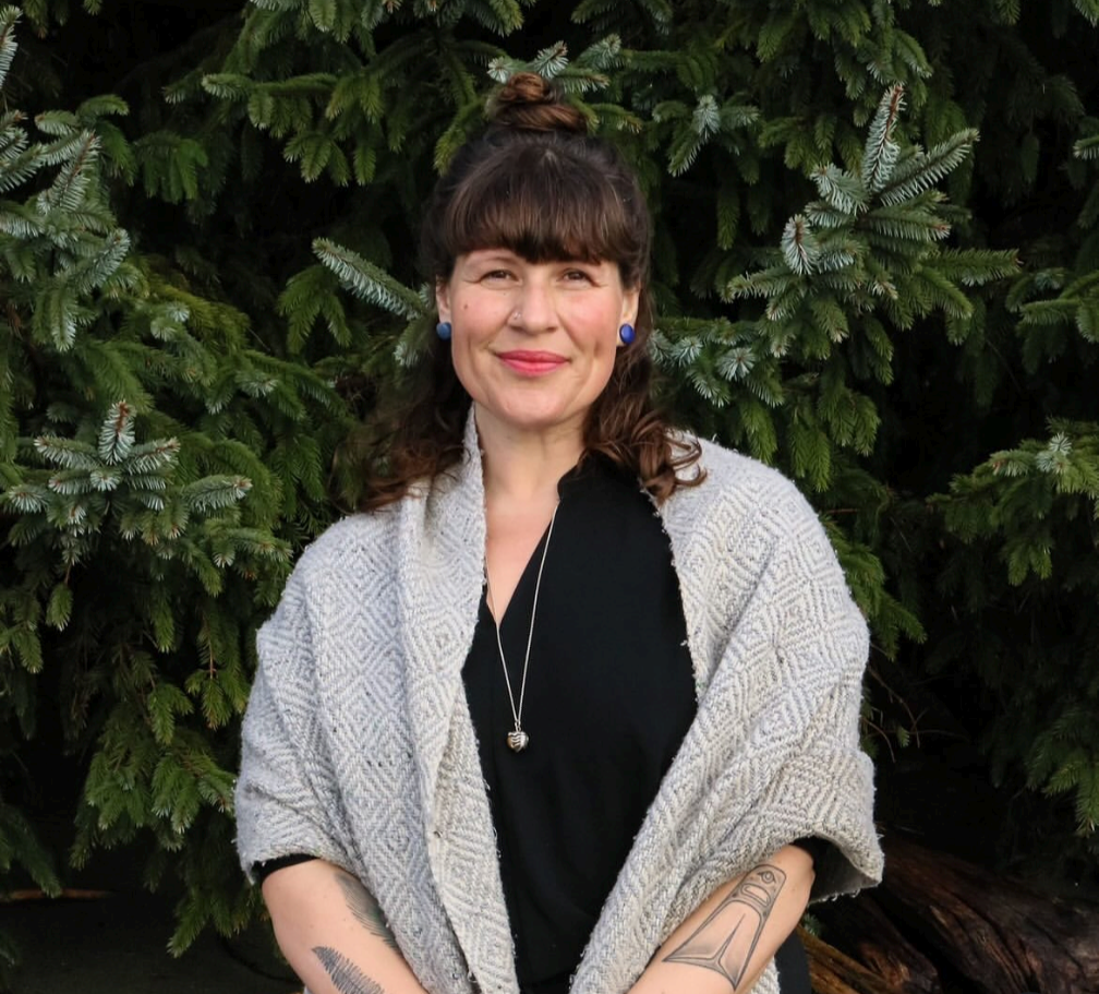 A woman (Melissa Hardy, Founder of Seeds of Vitality) with brown hair and bangs, wearing a black top, a gray patterned shawl, blue earrings, and a pendant necklace, standing outdoors in front of green pine trees, smiling at the camera.