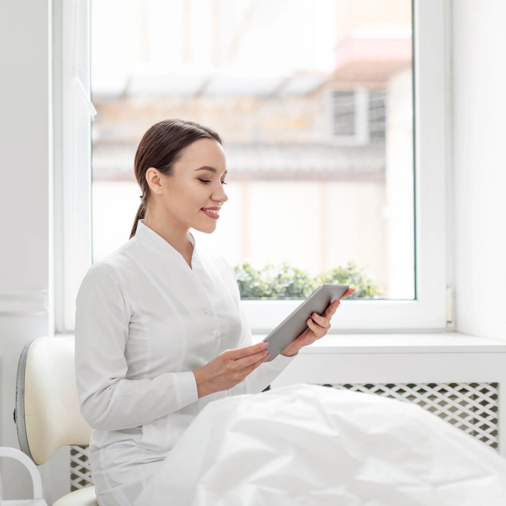 A young woman in a white medical coat sitting by a window and looking at a tablet.