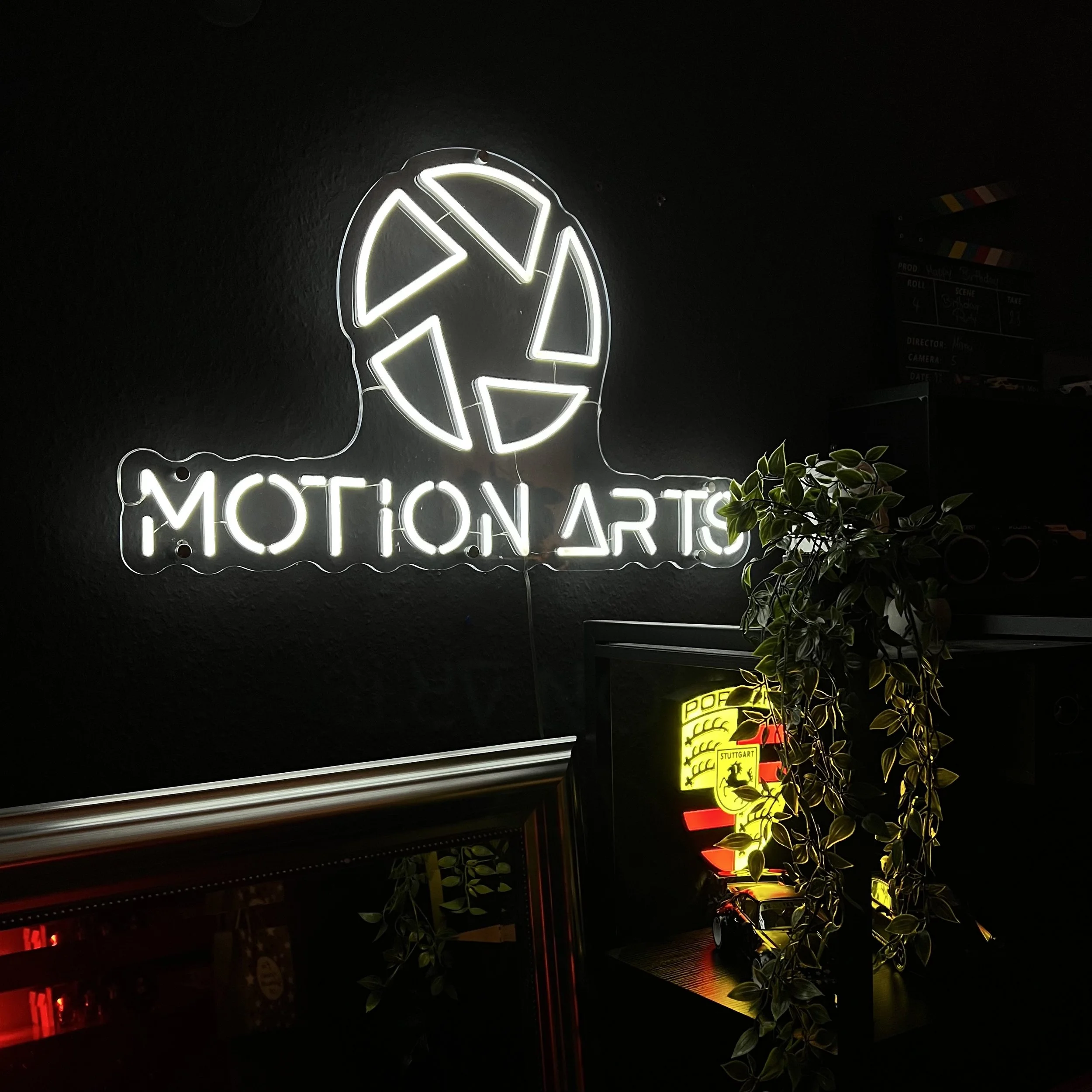 Leuchtschild mit dem Logo und dem Schriftzug 'MOTION ART' an einer schwarzen Wand, daneben eine Pflanze und ein weiteres leuchtendes Schild mit Porsche-Logo.