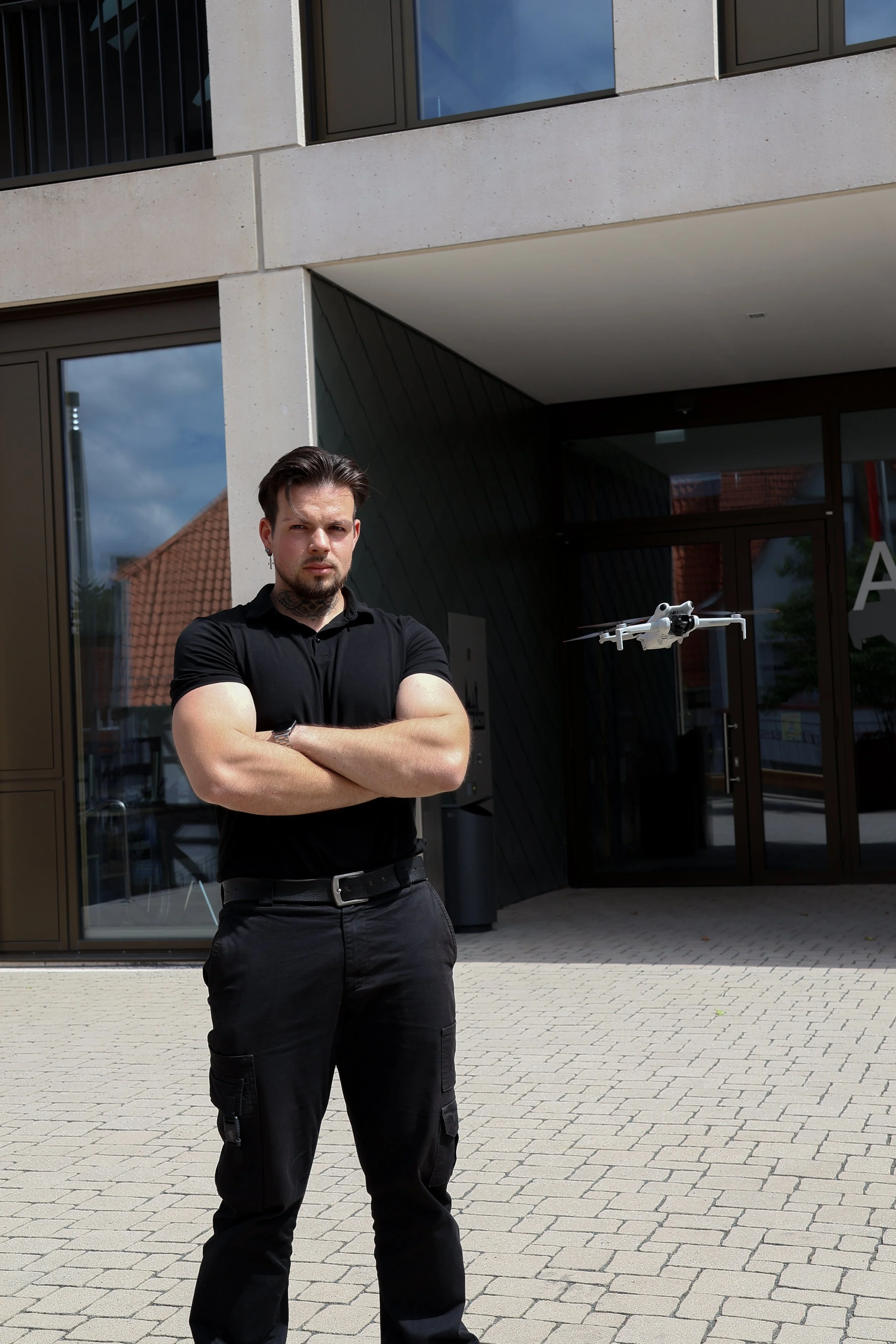 Ein Mann steht mit verschränkten Armen vor einem modernen Gebäude mit großen Fenstern und einem Drohnenflugzeug im Hintergrund.