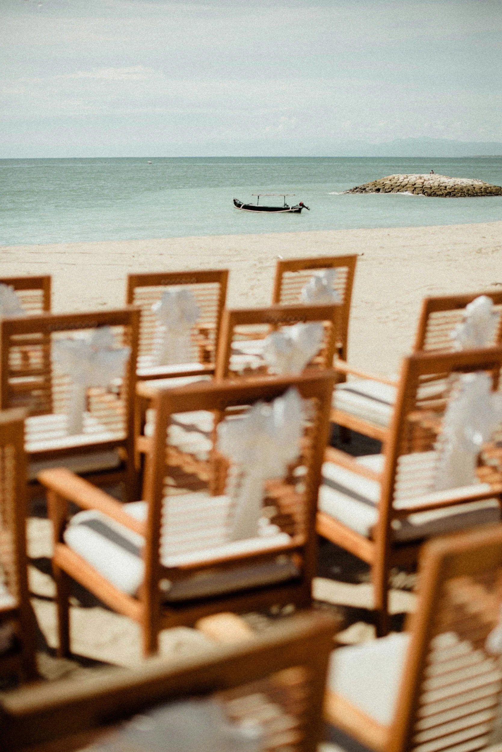 Wooden chairs on a sandy beach with a boat, a rocky pier, and calm water in the background.