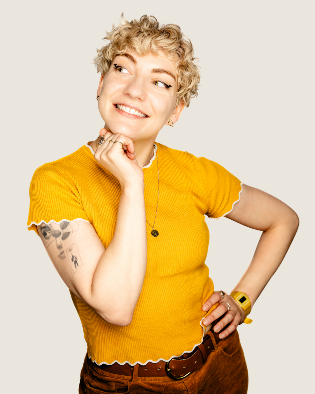 A woman with short, curly blonde hair wearing a yellow ribbed shirt with white trim, brown pants, and a yellow watch. She has tattoos on her left arm, multiple rings, and a necklace. She is smiling and looking to the side with her left hand on her hip and her right hand resting on her chin.