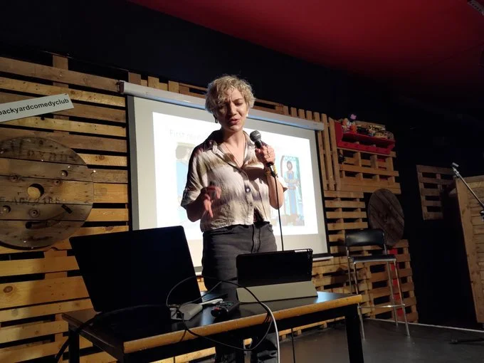 A woman standing on stage giving a presentation at a comedy club, holding a microphone with a projected screen behind her, in a rustic setting with wooden pallet decor on the walls.