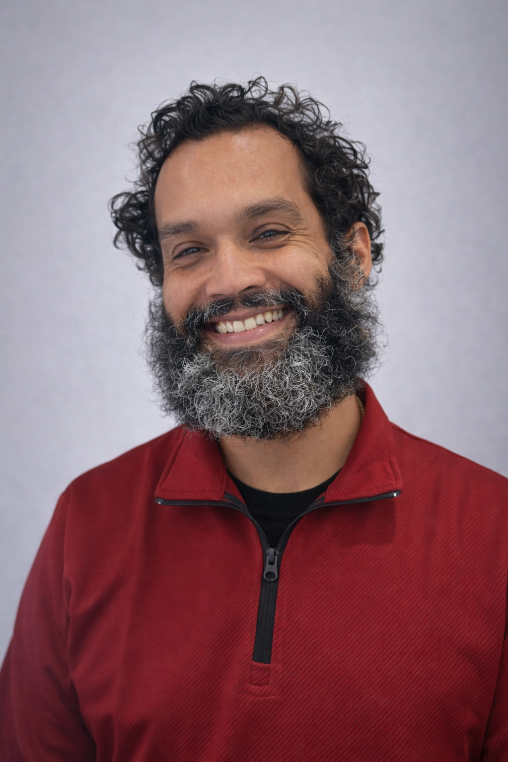 A smiling man with curly hair and a full beard, wearing a red zip-up jacket, against a light-colored background.