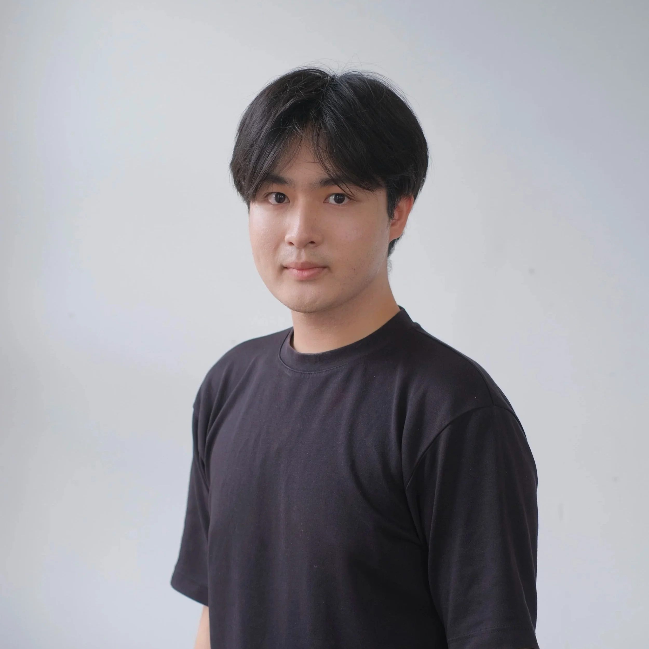 A young man with black hair wearing a black T-shirt, standing against a plain white wall.