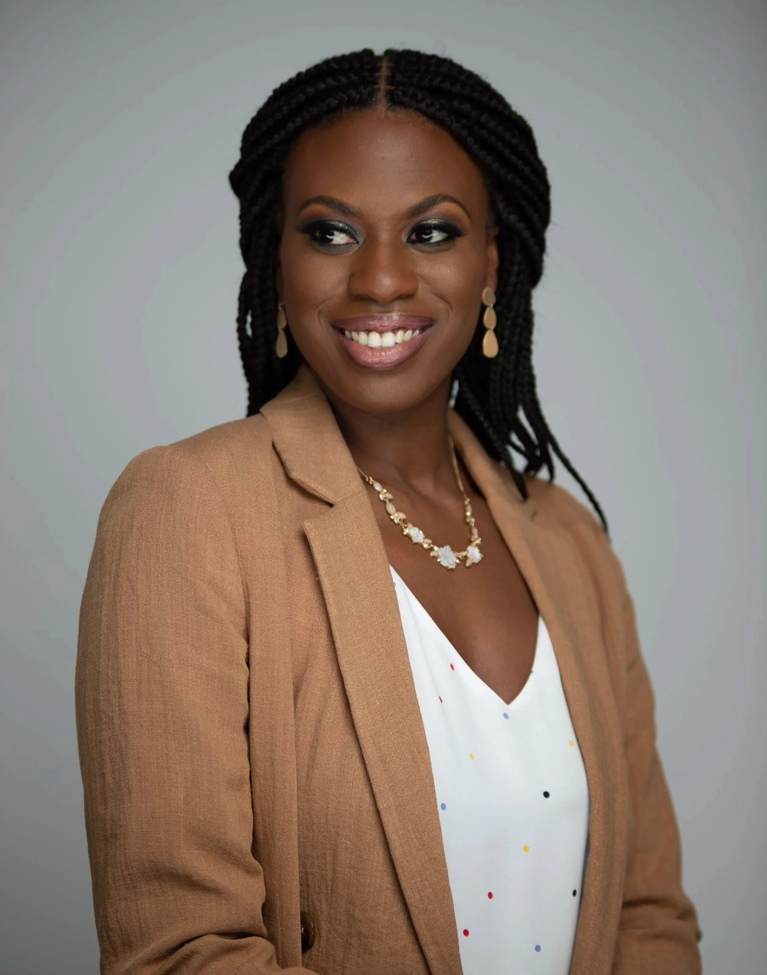 A smiling woman with braided hair, wearing a tan blazer, white blouse with colorful polka dots, gold earrings, and a necklace, presenting a professional appearance.