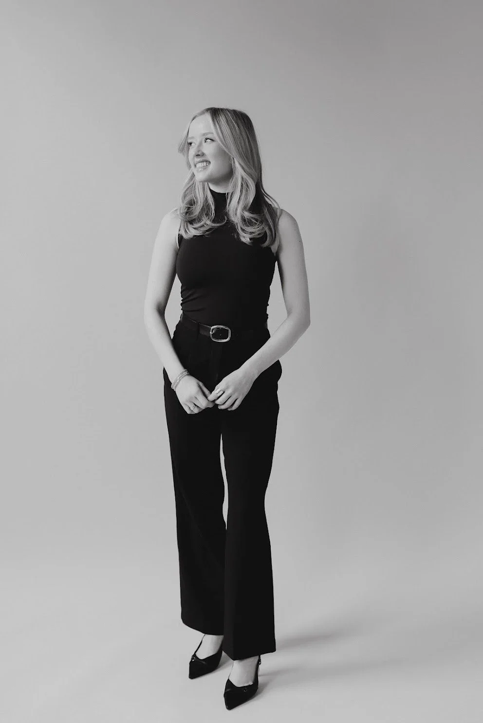 A woman in black sleeveless top and black pants standing with her hands clasped, smiling and looking to her right against a plain light background.