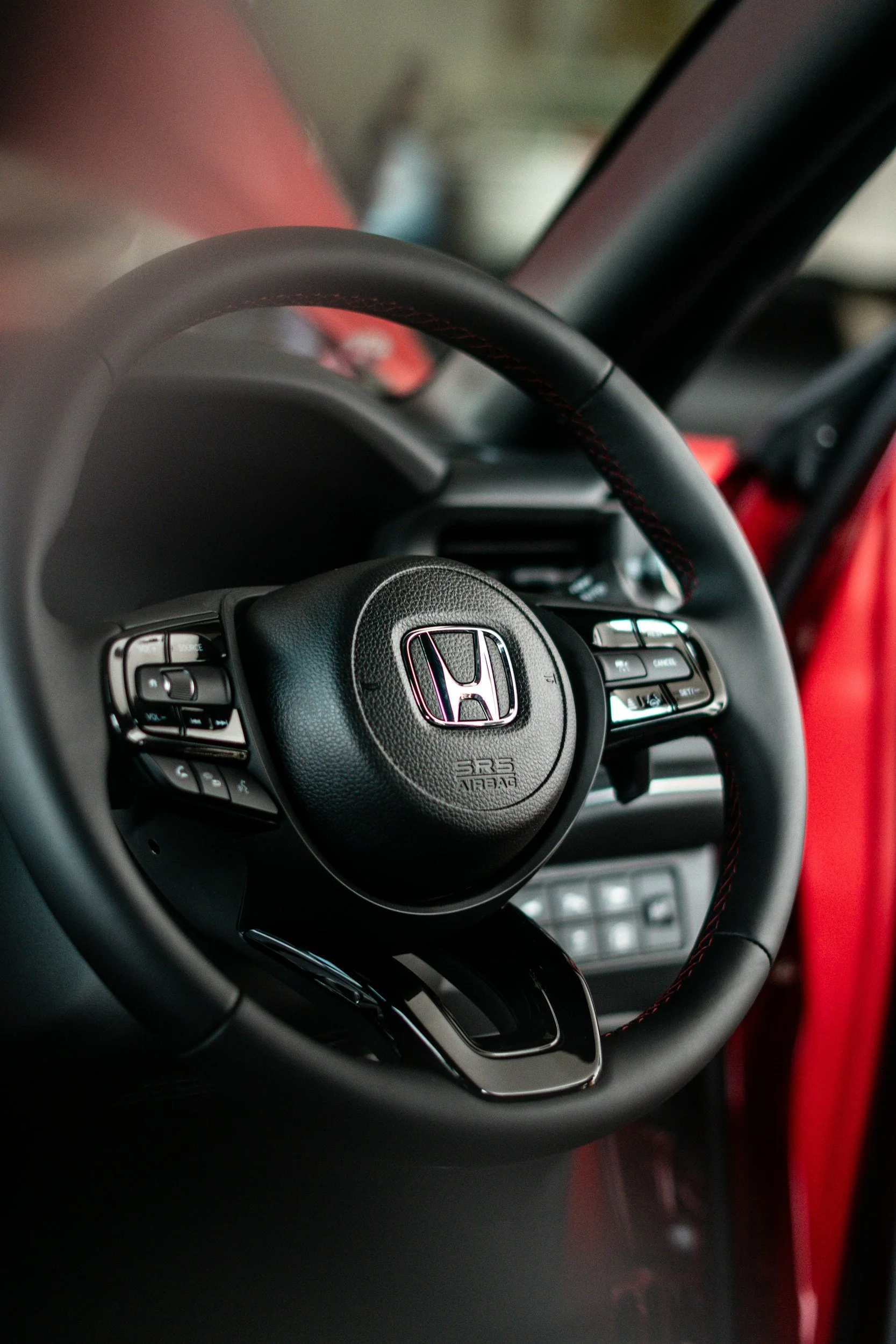 Close-up of a black Honda steering wheel with control buttons, interior view of a car with part of the dashboard and red interior visible.