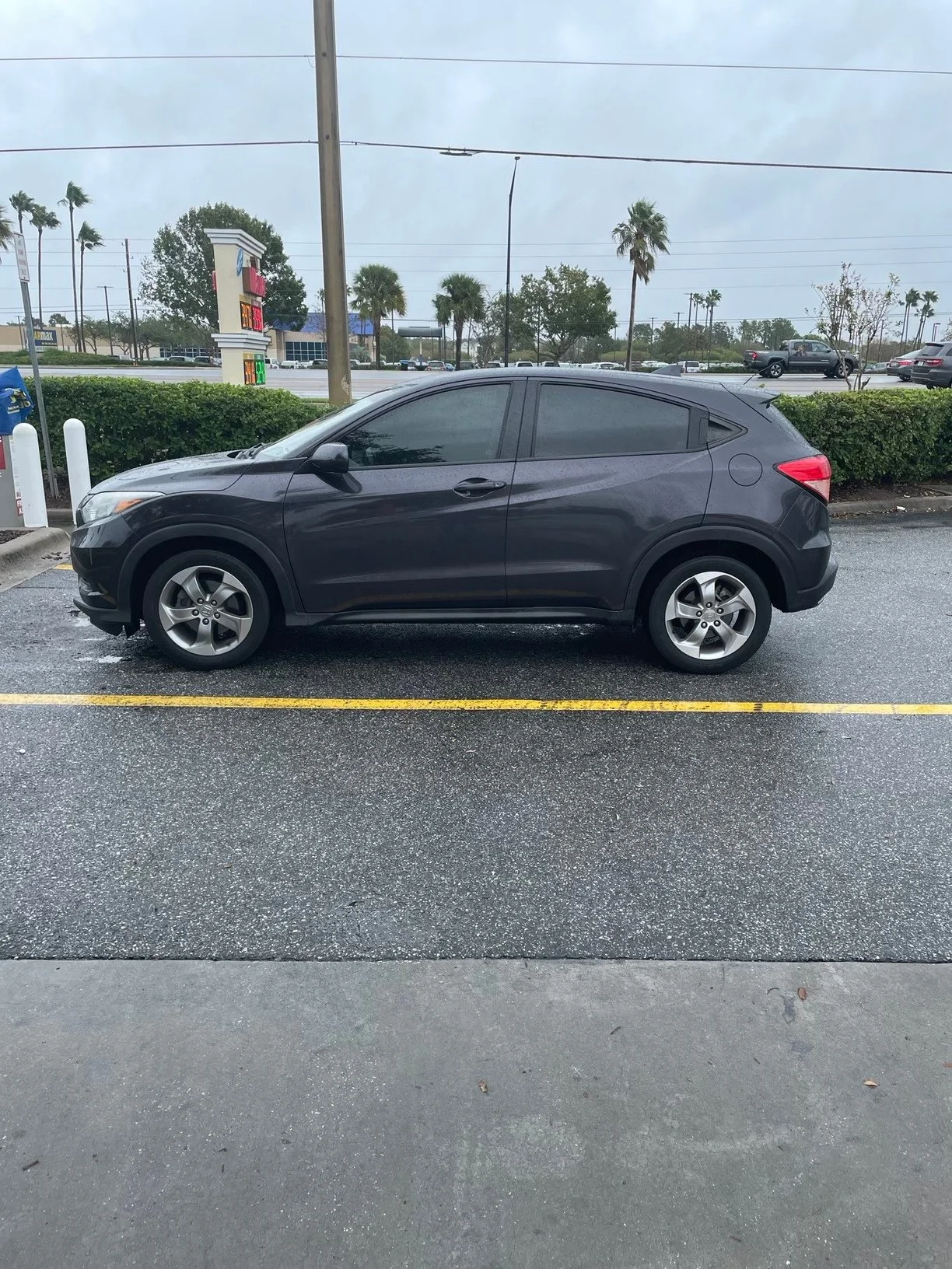 A gray Honda HR-V parked in a parking lot with trees, palm trees, shopping center, and cloudy sky in the background on a rainy day.