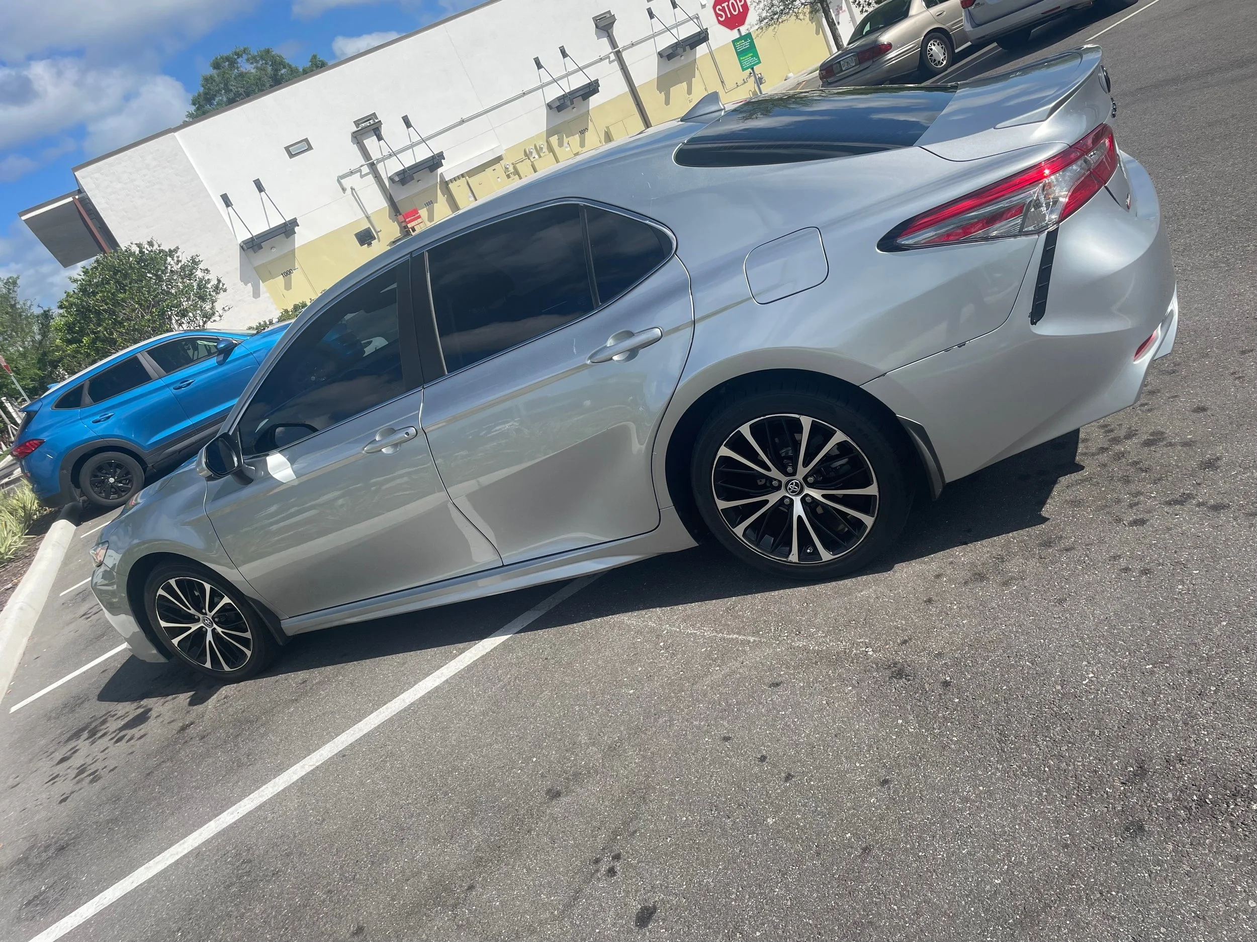 Silver sedan parked in a parking lot with a blue SUV parked nearby and a white building with black awnings in the background.